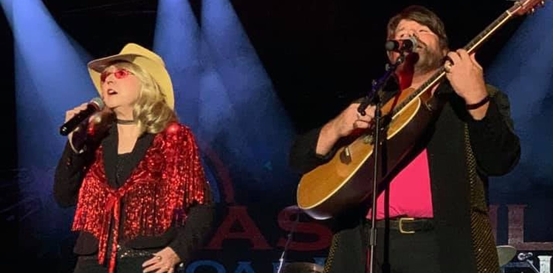 Two people on stage performing; one sings and one plays guitar. Stage lights and a Texas Roadhouse sign.