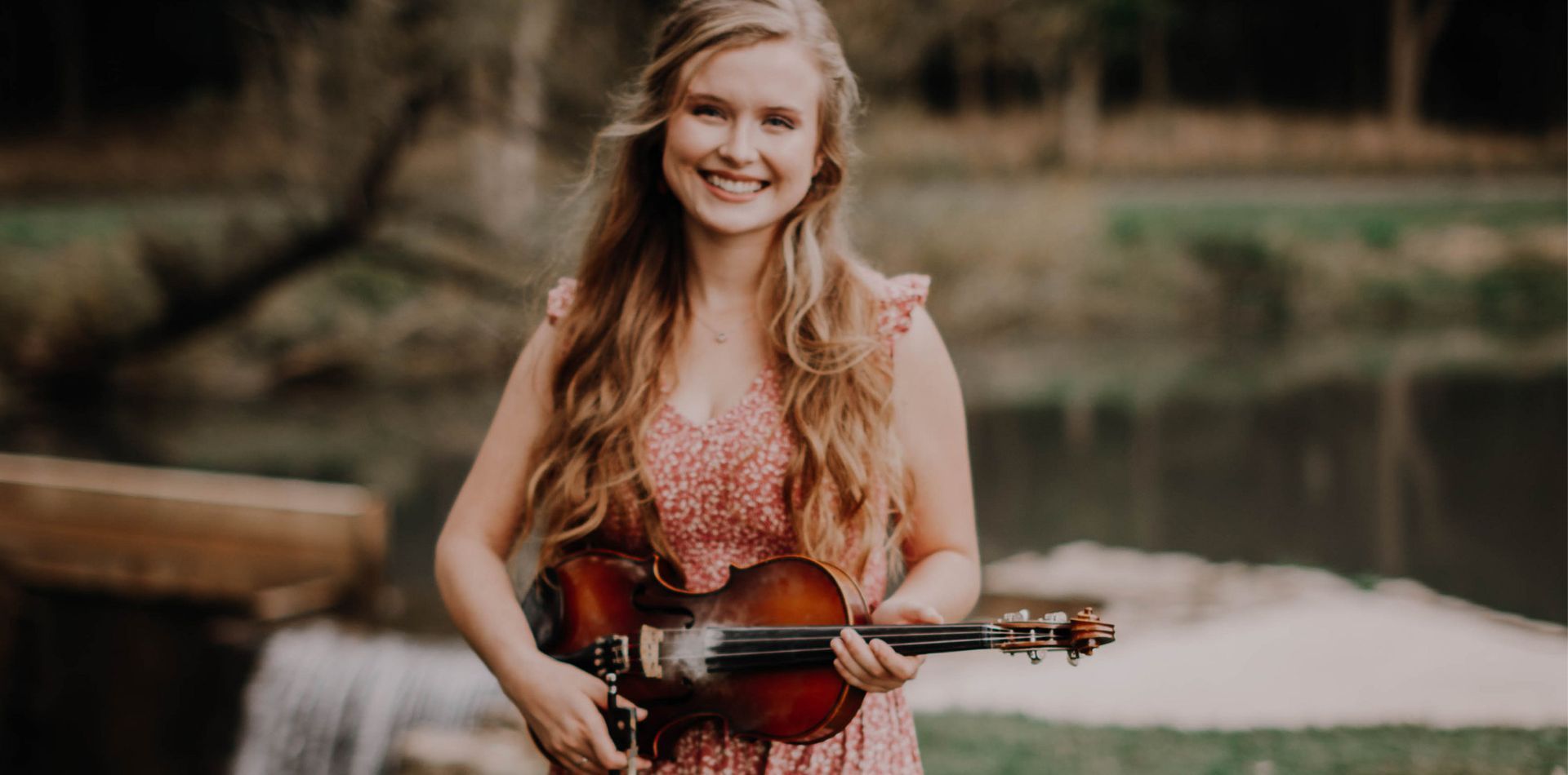 A woman in a pink dress is holding a violin in front of a waterfall.