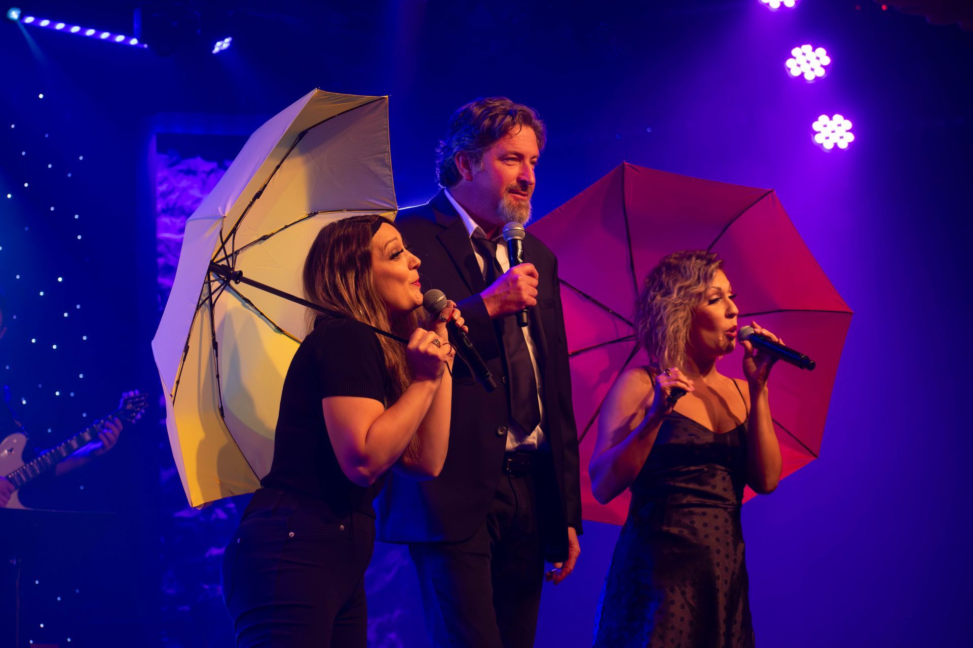A man and two women are singing into microphones while holding umbrellas on a stage.