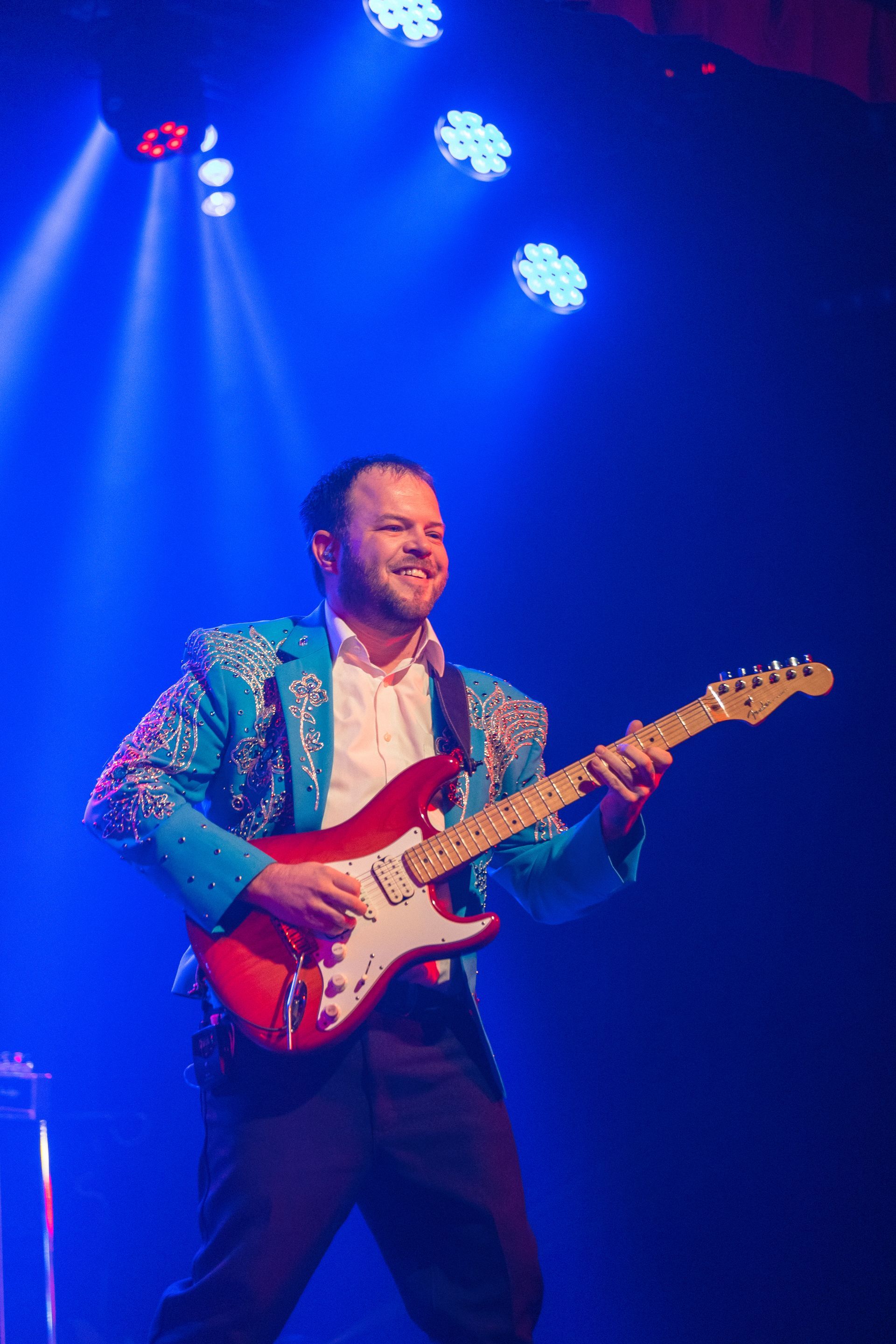 A man in a blue jacket is playing an electric guitar on a stage.