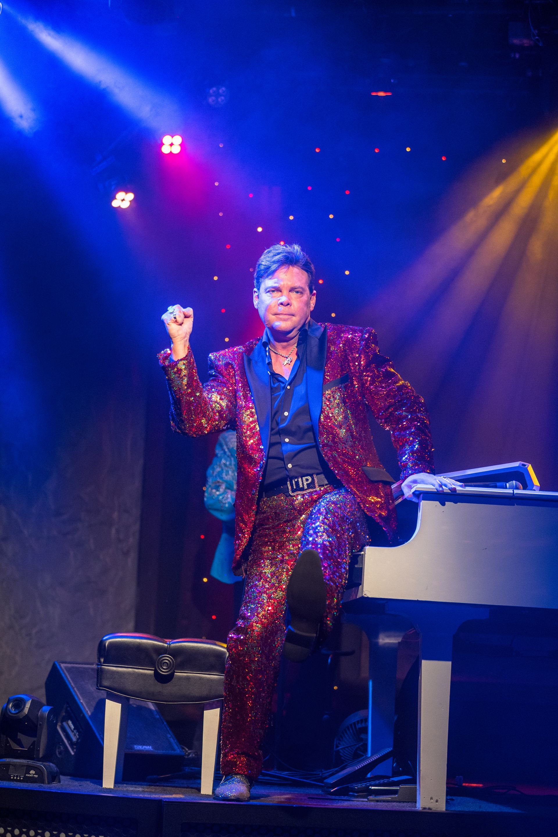 A man in a red sequined suit is sitting at a white piano on a stage.