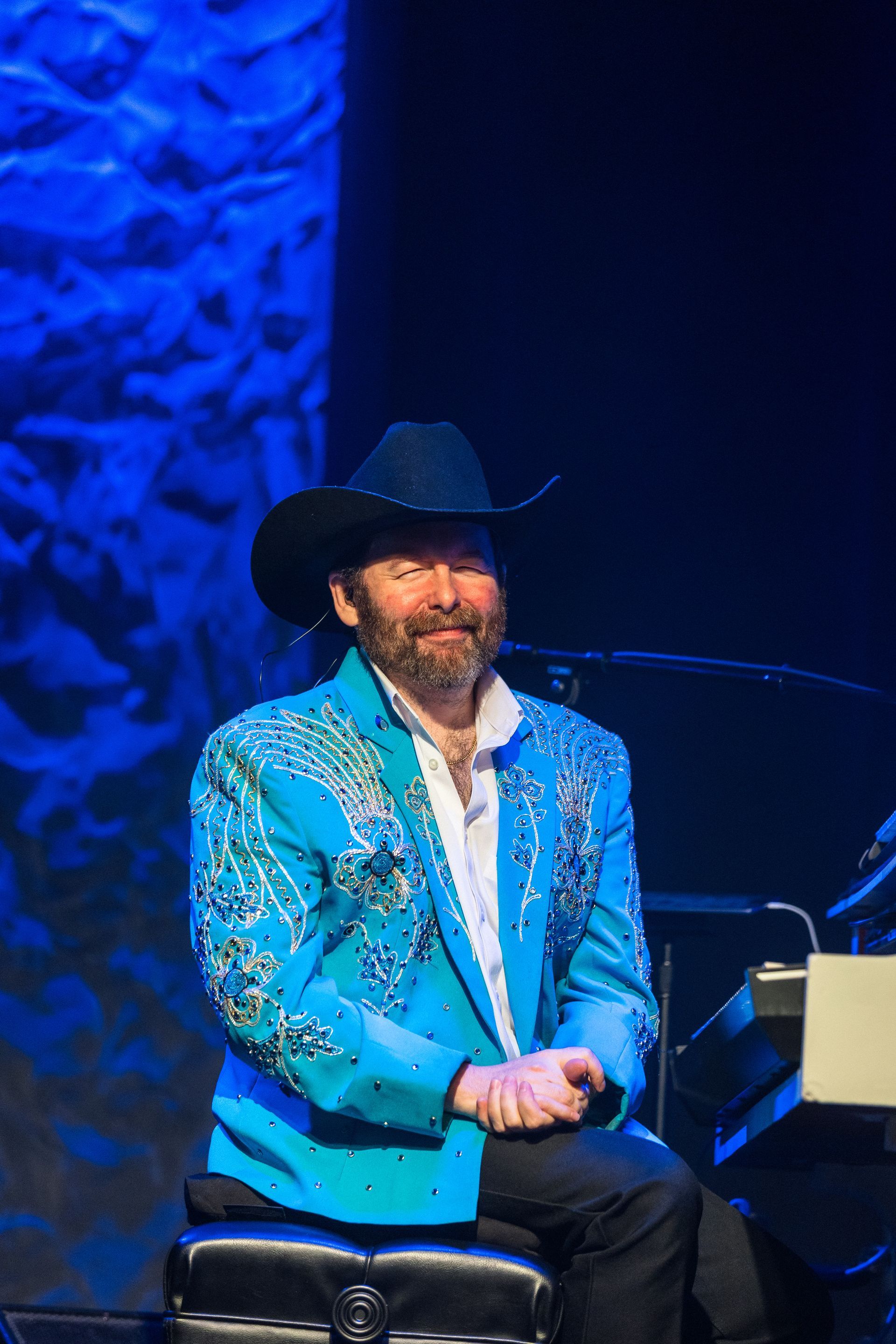 A man in a blue jacket and cowboy hat is sitting at a piano on a stage.