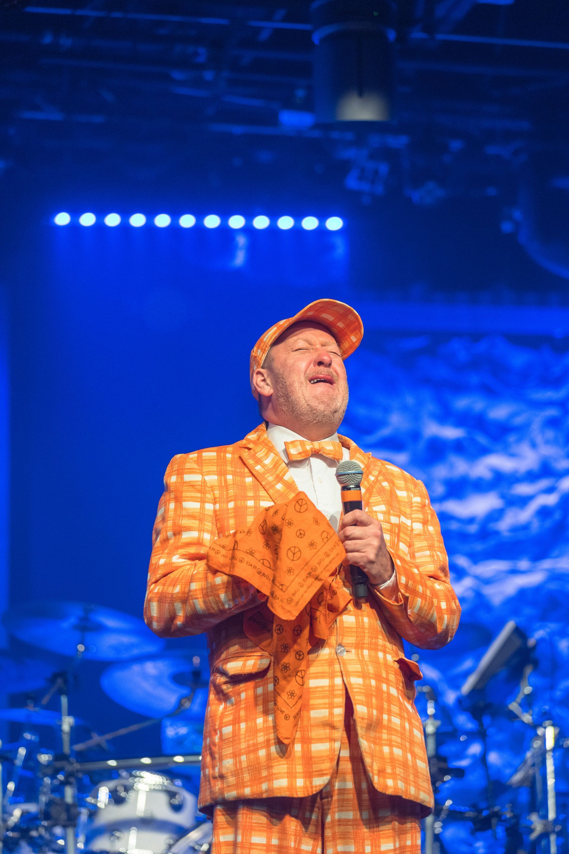 A man in an orange suit is singing into a microphone on a stage.