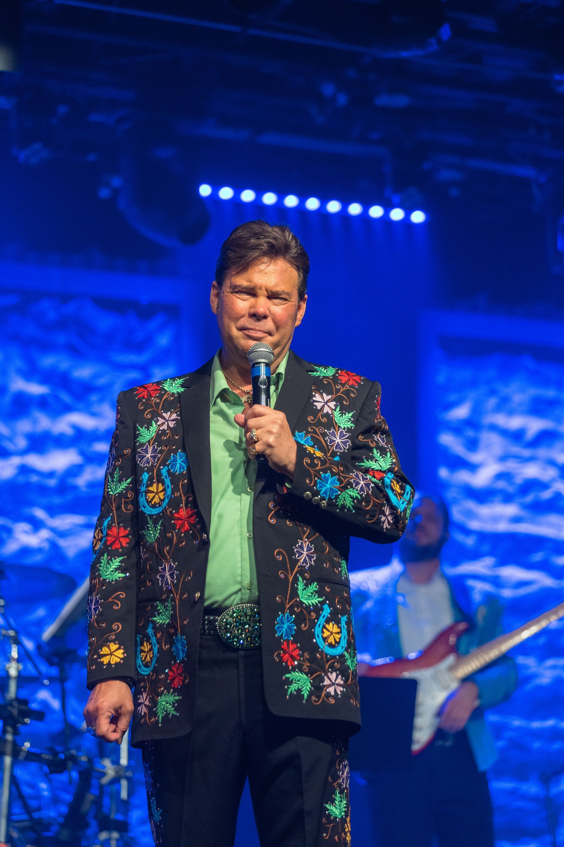 A man in a floral jacket is holding a microphone on a stage