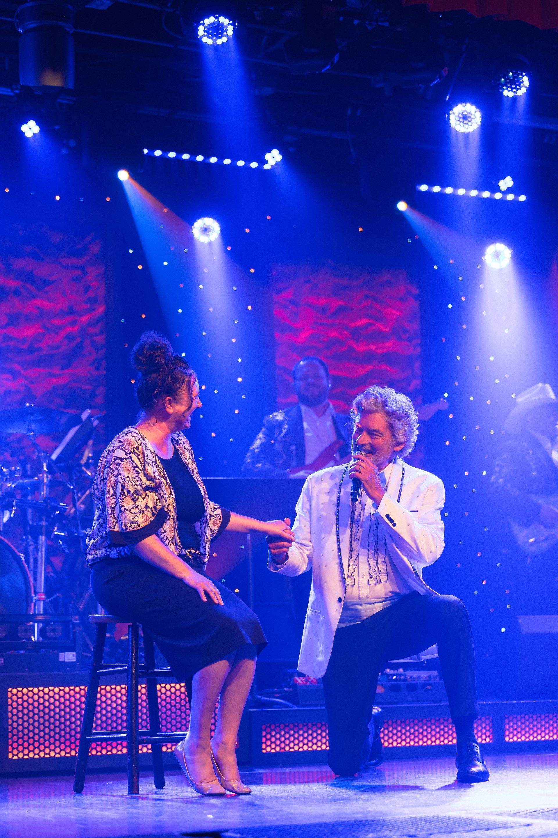 A man is singing into a microphone while a woman sits on a stool next to him on a stage.