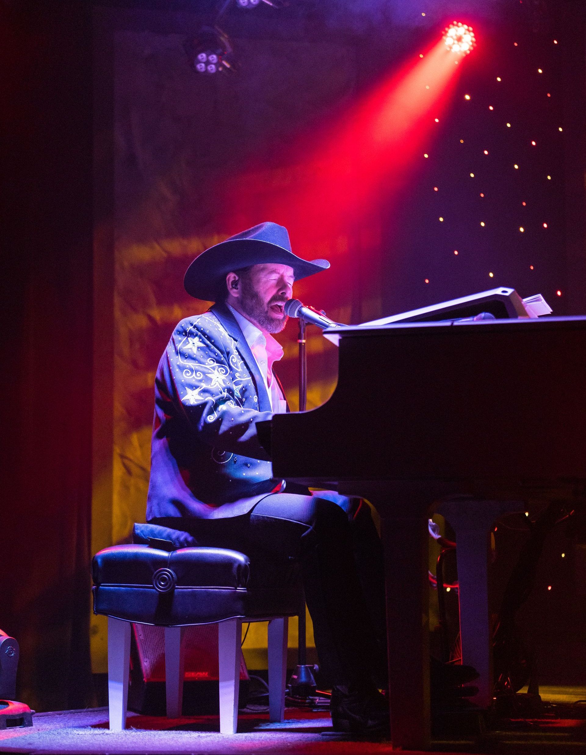 A man in a cowboy hat is singing into a microphone while playing a piano
