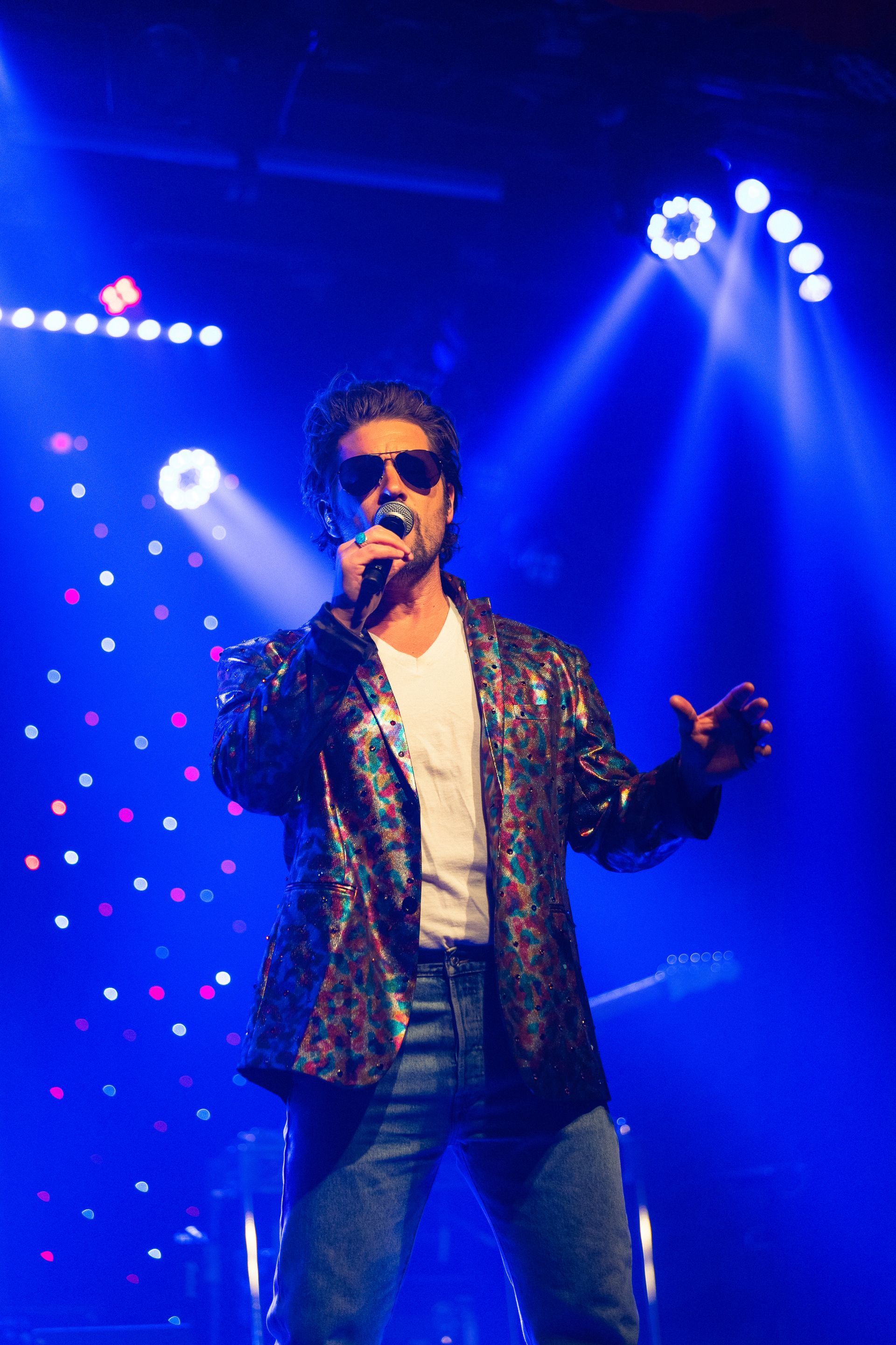 A man in a sequined jacket is singing into a microphone on a stage.