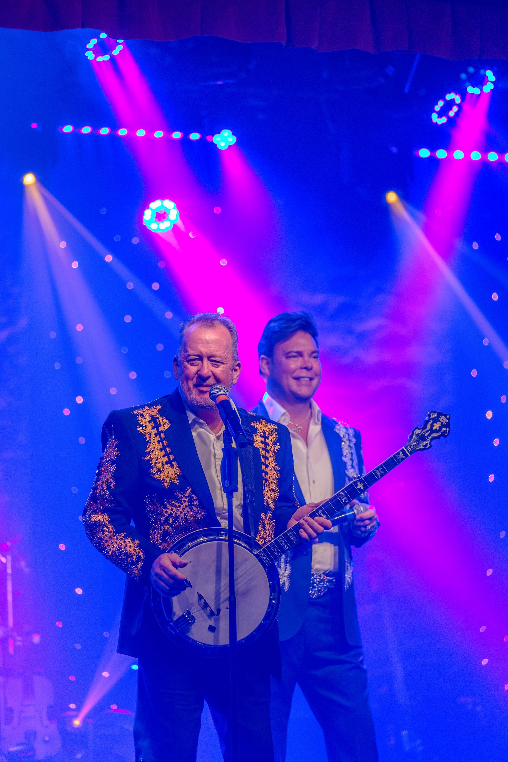 Two men are playing banjos and singing into microphones on a stage.