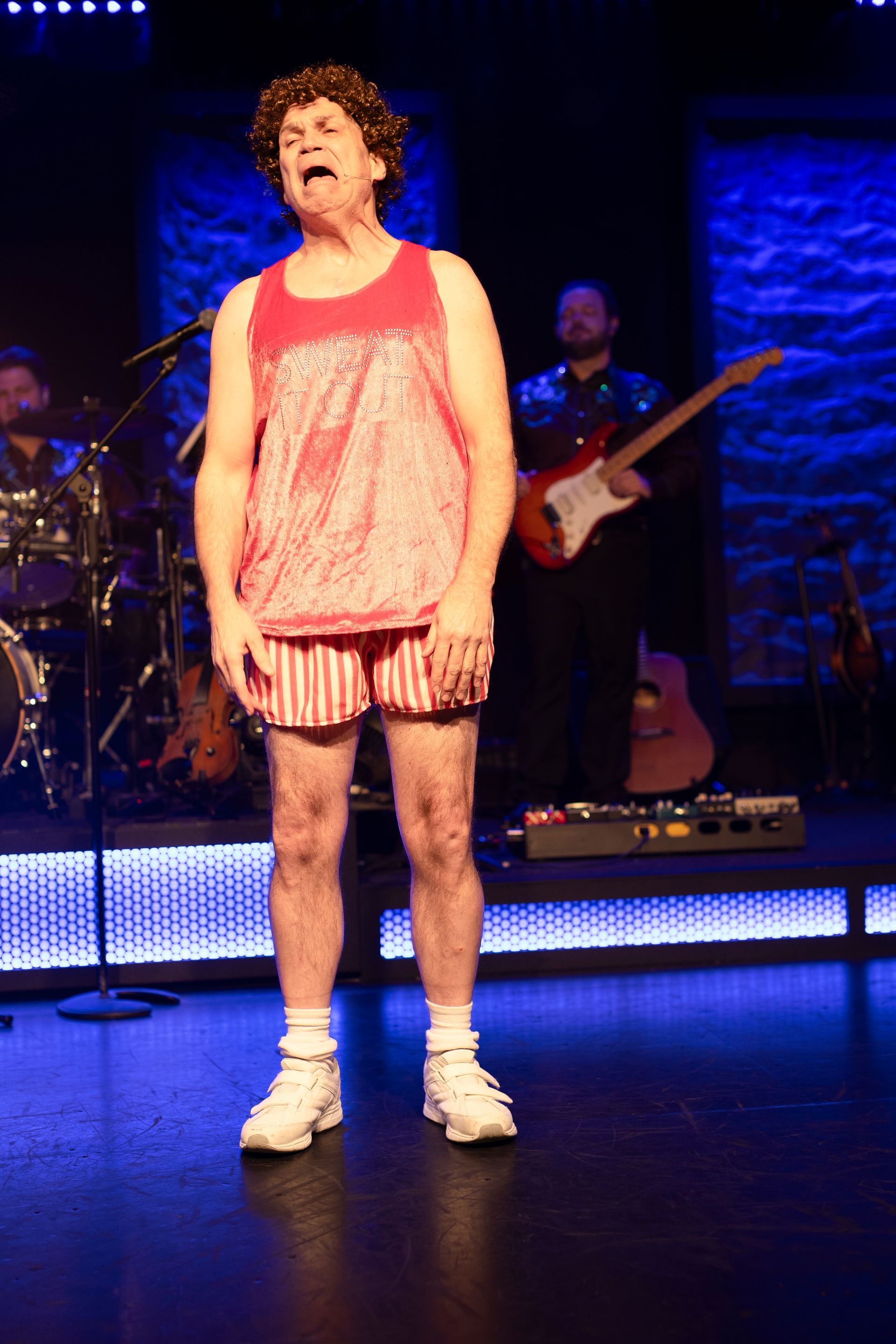 A man in a red tank top and shorts is singing into a microphone on a stage.