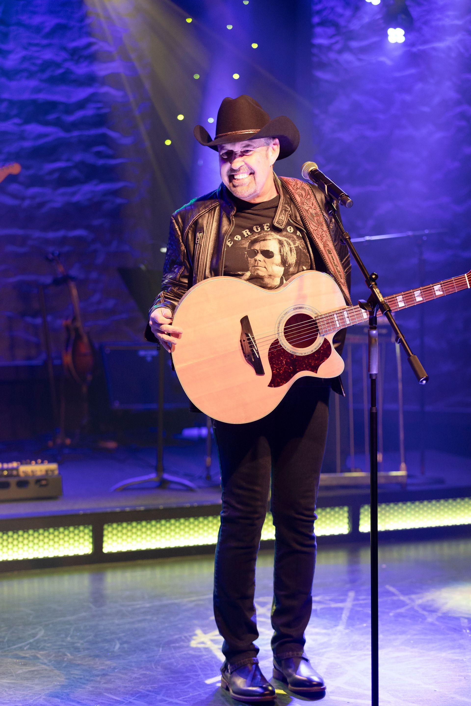 A man in a cowboy hat is holding a guitar and singing into a microphone on a stage.