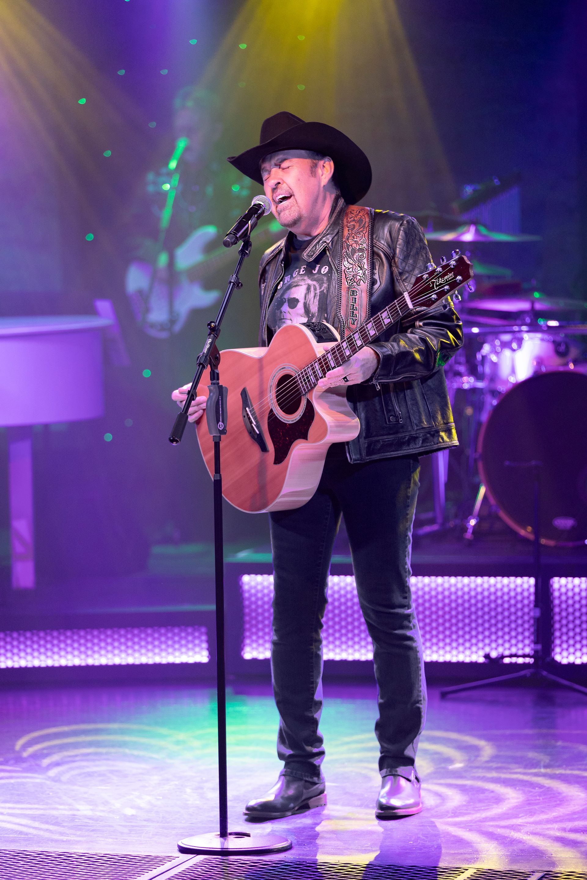 A man in a cowboy hat is playing an acoustic guitar and singing into a microphone on a stage.