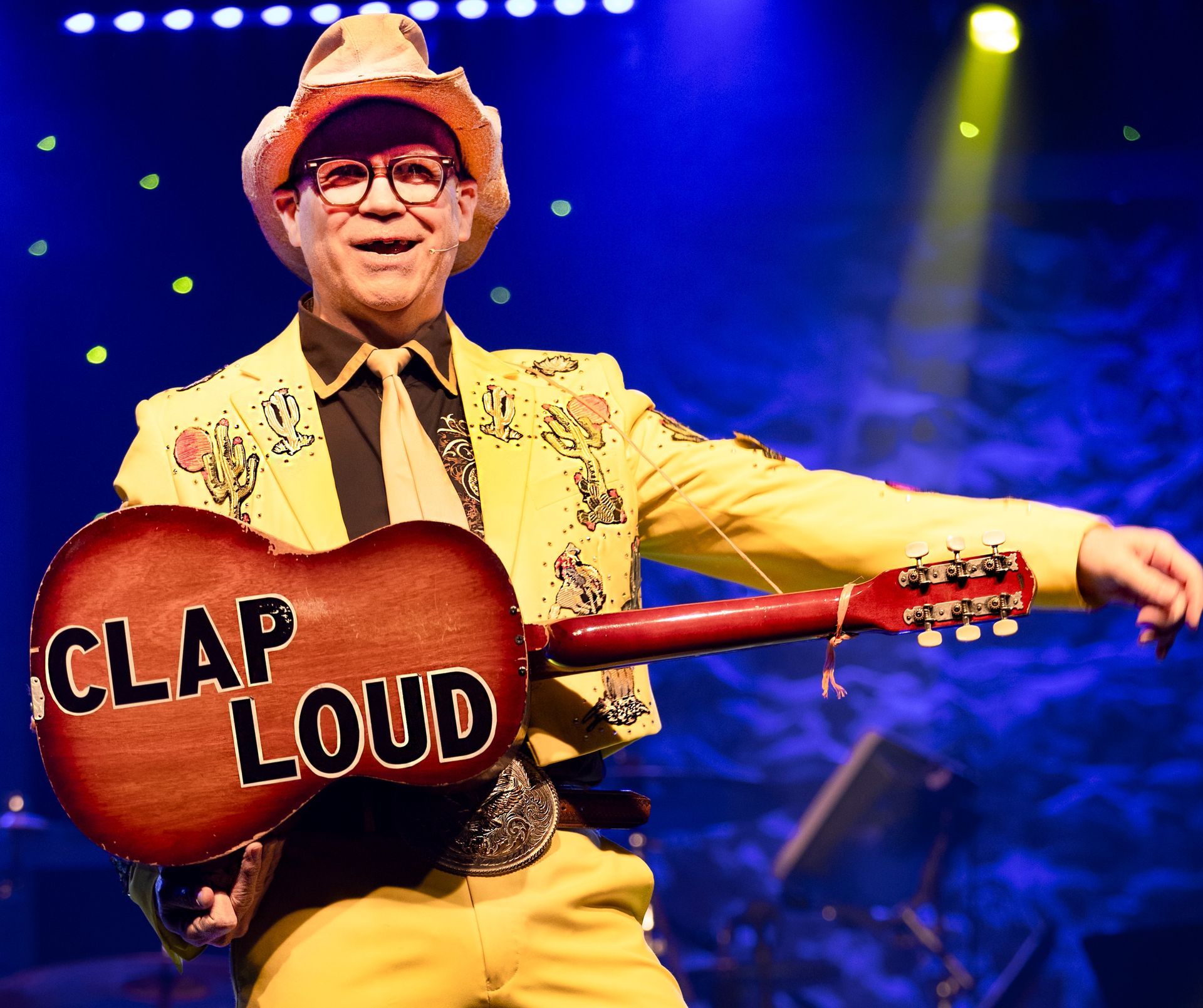 A man in a yellow suit is holding a guitar that says clap loud