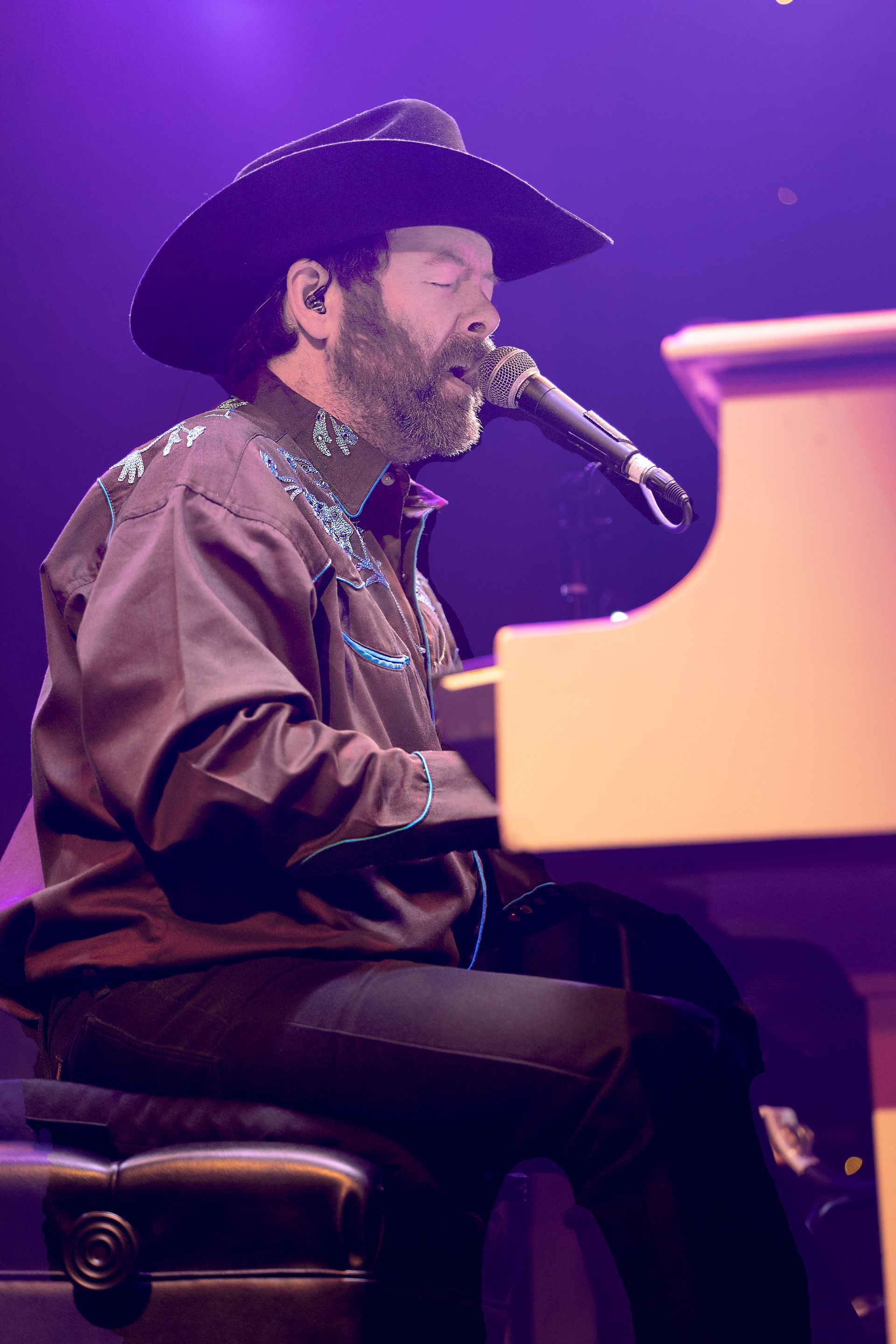 A man in a cowboy hat is singing into a microphone while playing a piano.