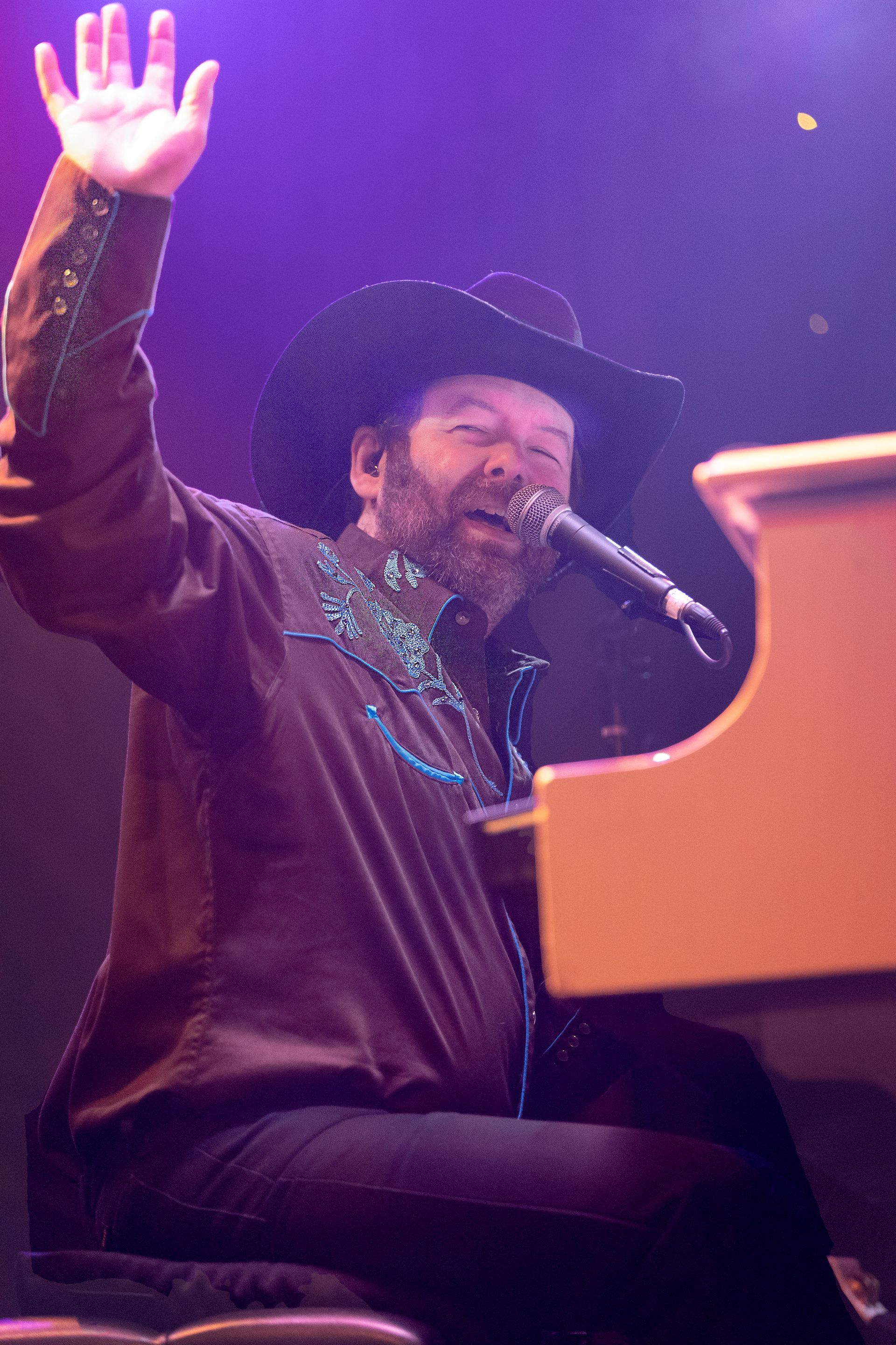 A man in a cowboy hat is singing into a microphone while playing a piano