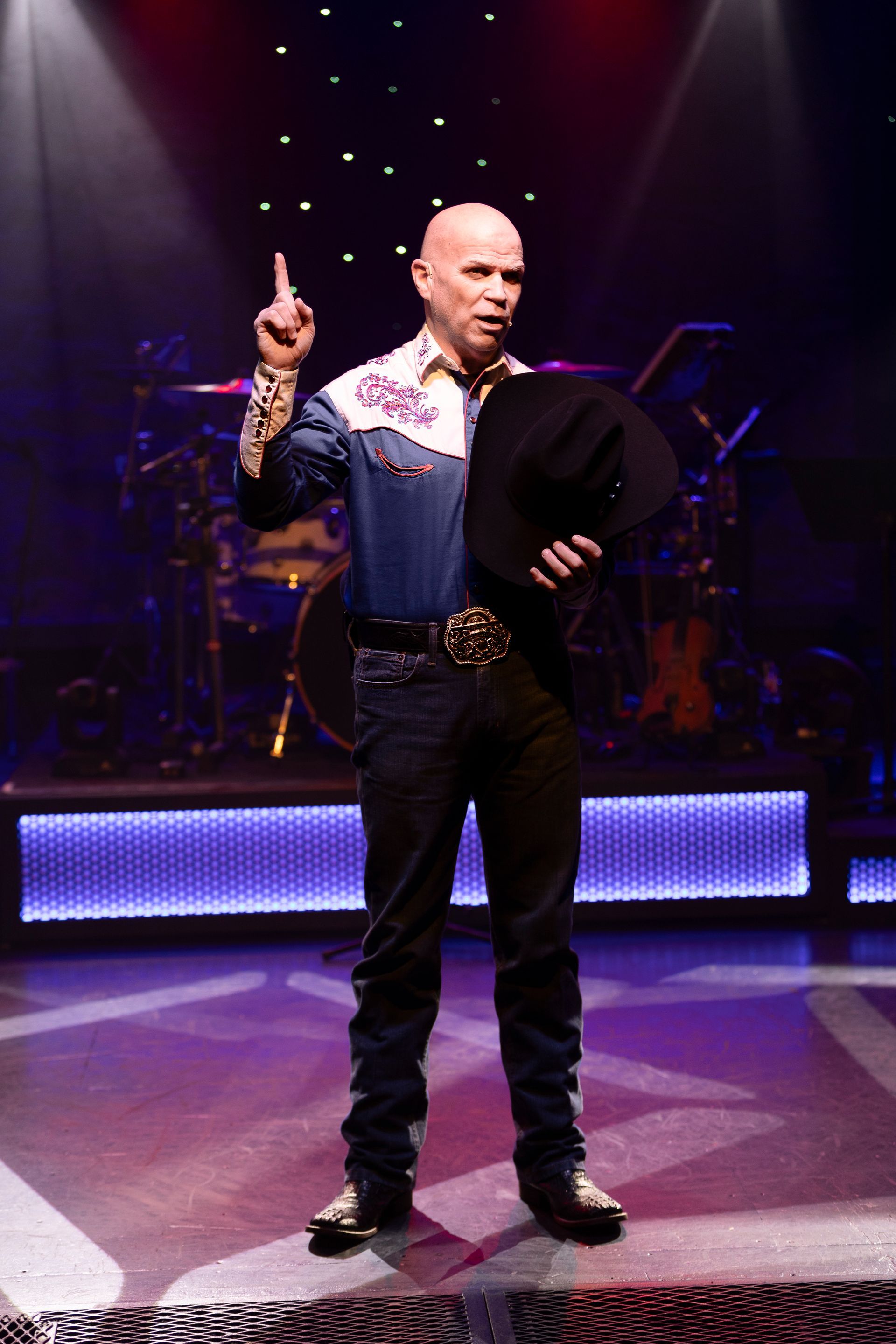 A bald man is standing on a stage holding a cowboy hat and pointing up.