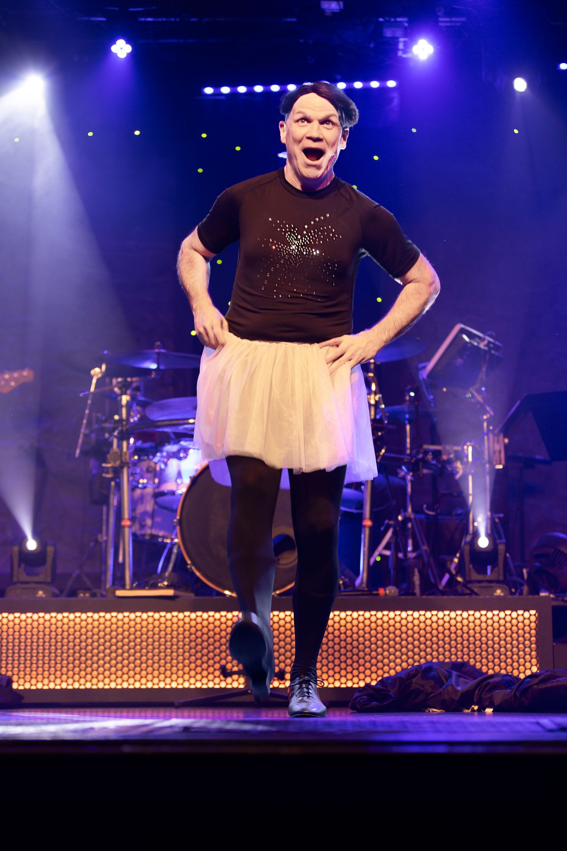 A man in a black shirt and white skirt is dancing on a stage
