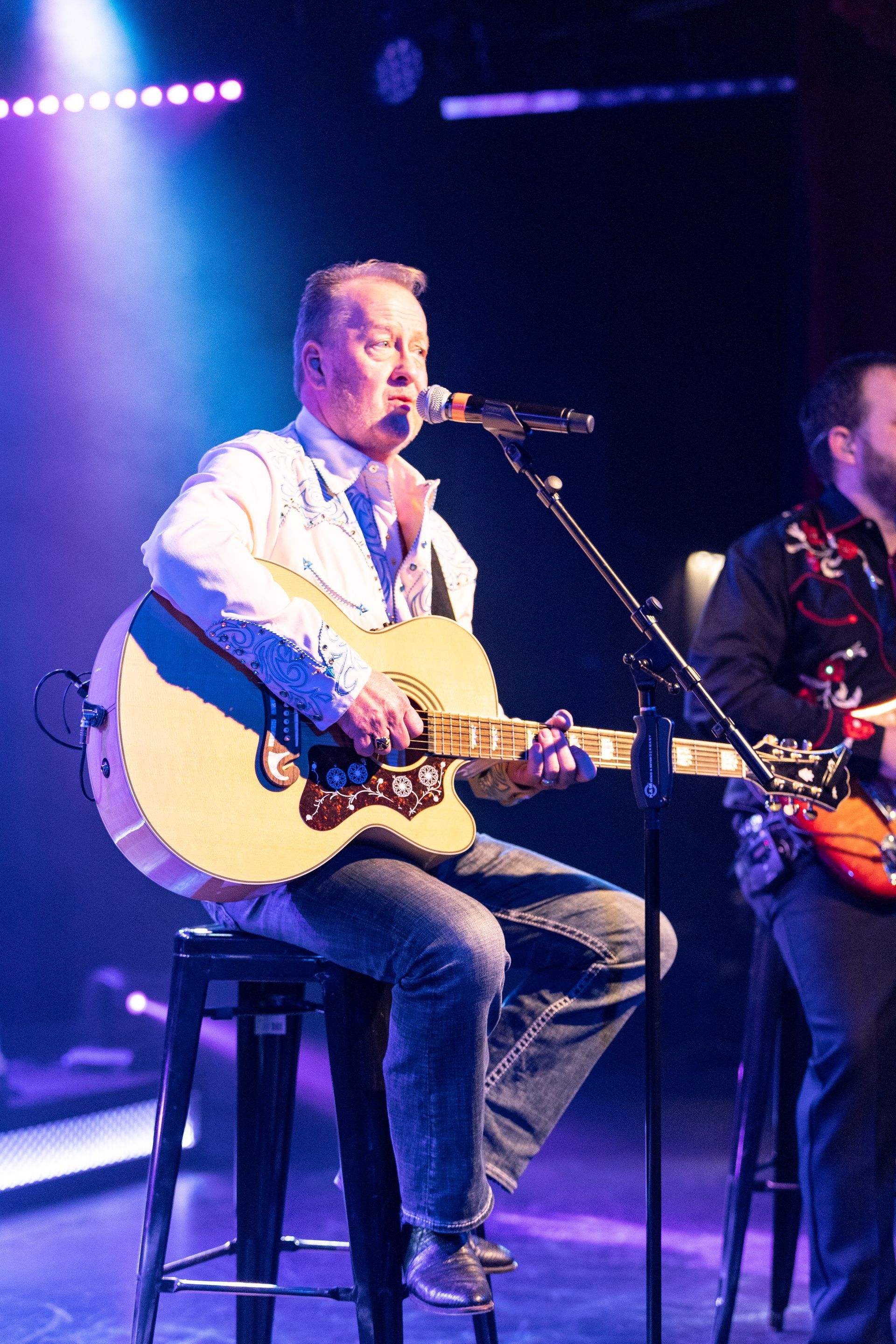 A man is sitting on a stool playing a guitar and singing into a microphone.