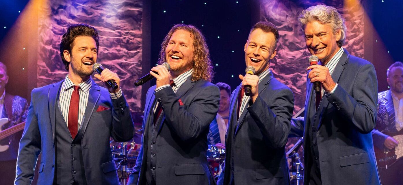 Four men in suits singing into microphones on a stage, smiling. Band in background.