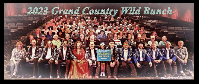 A large group of people are sitting in front of a sign that says grand country wild bunch.