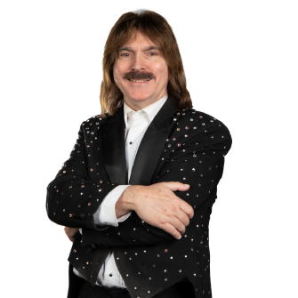 A man with long hair and a mustache is wearing a black tuxedo