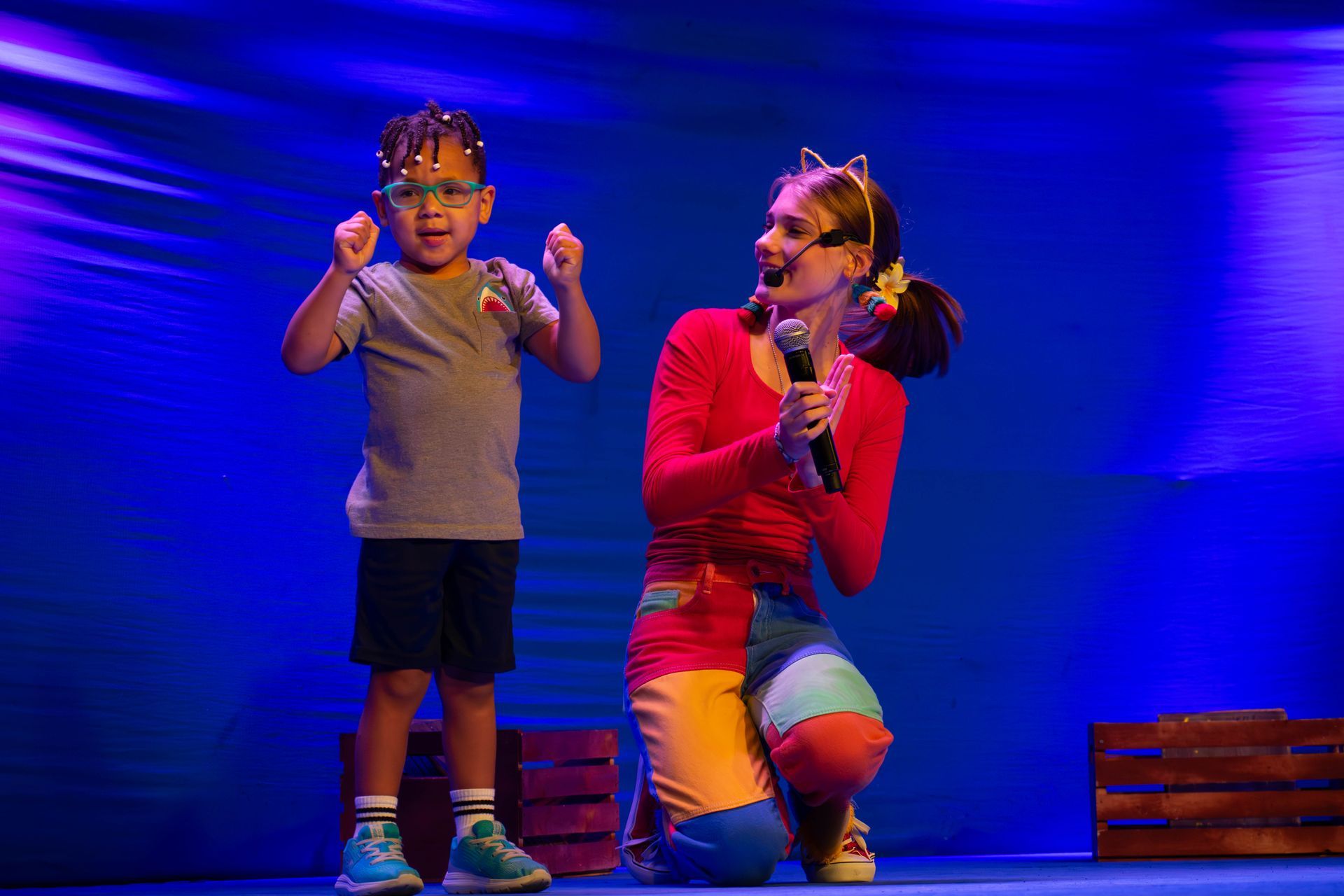 A woman is singing into a microphone while kneeling next to a little boy on a stage.