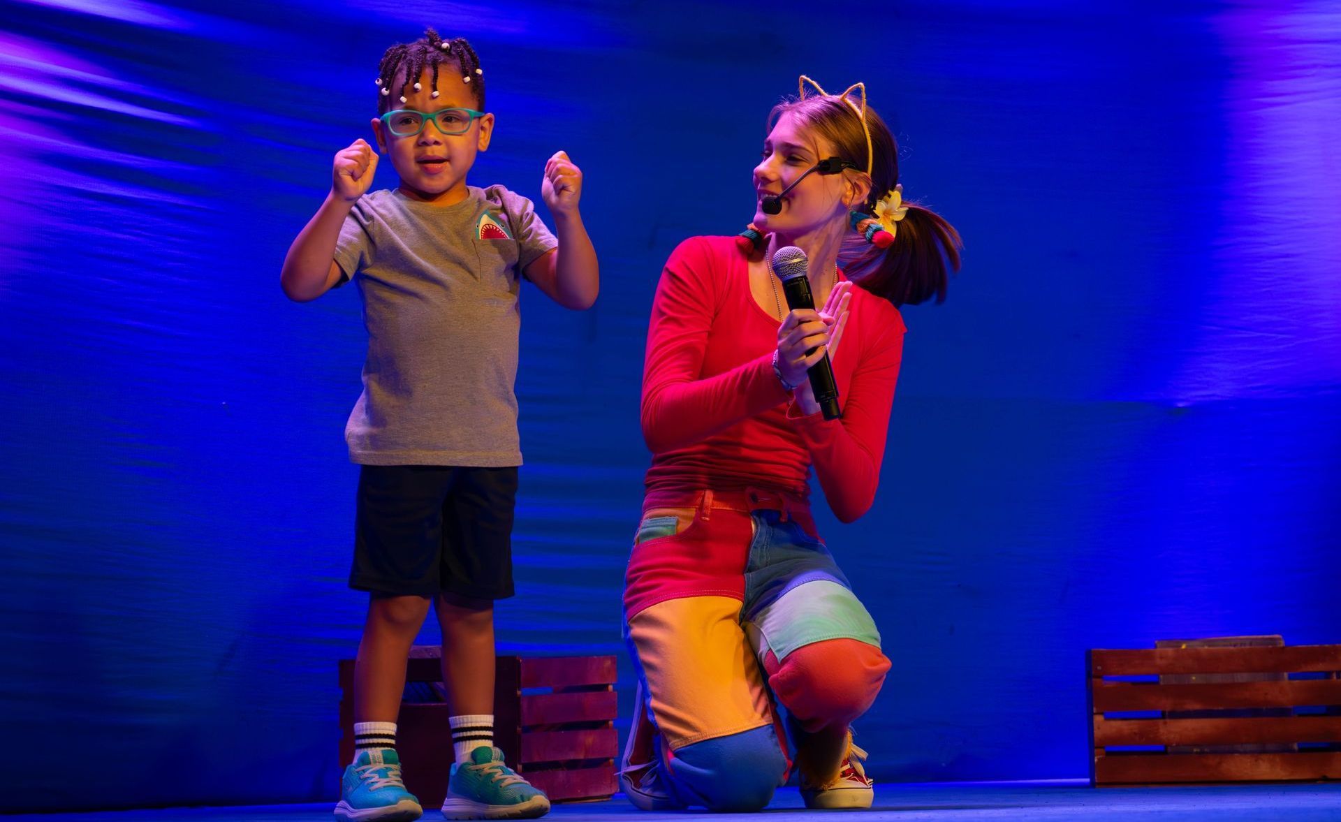 A woman is singing into a microphone while kneeling next to a little boy on a stage.