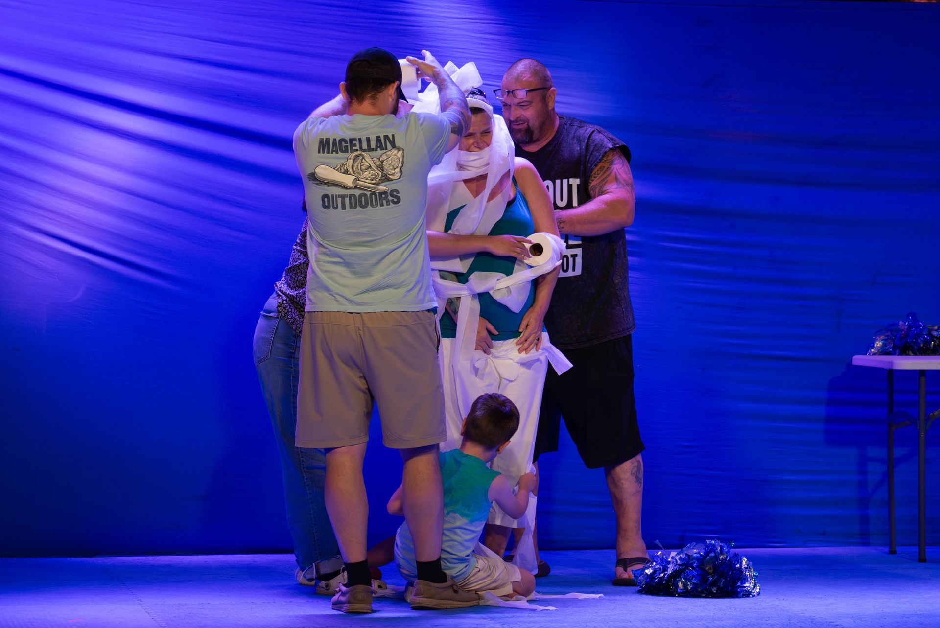 A group of people are standing around a woman wrapped in toilet paper.
