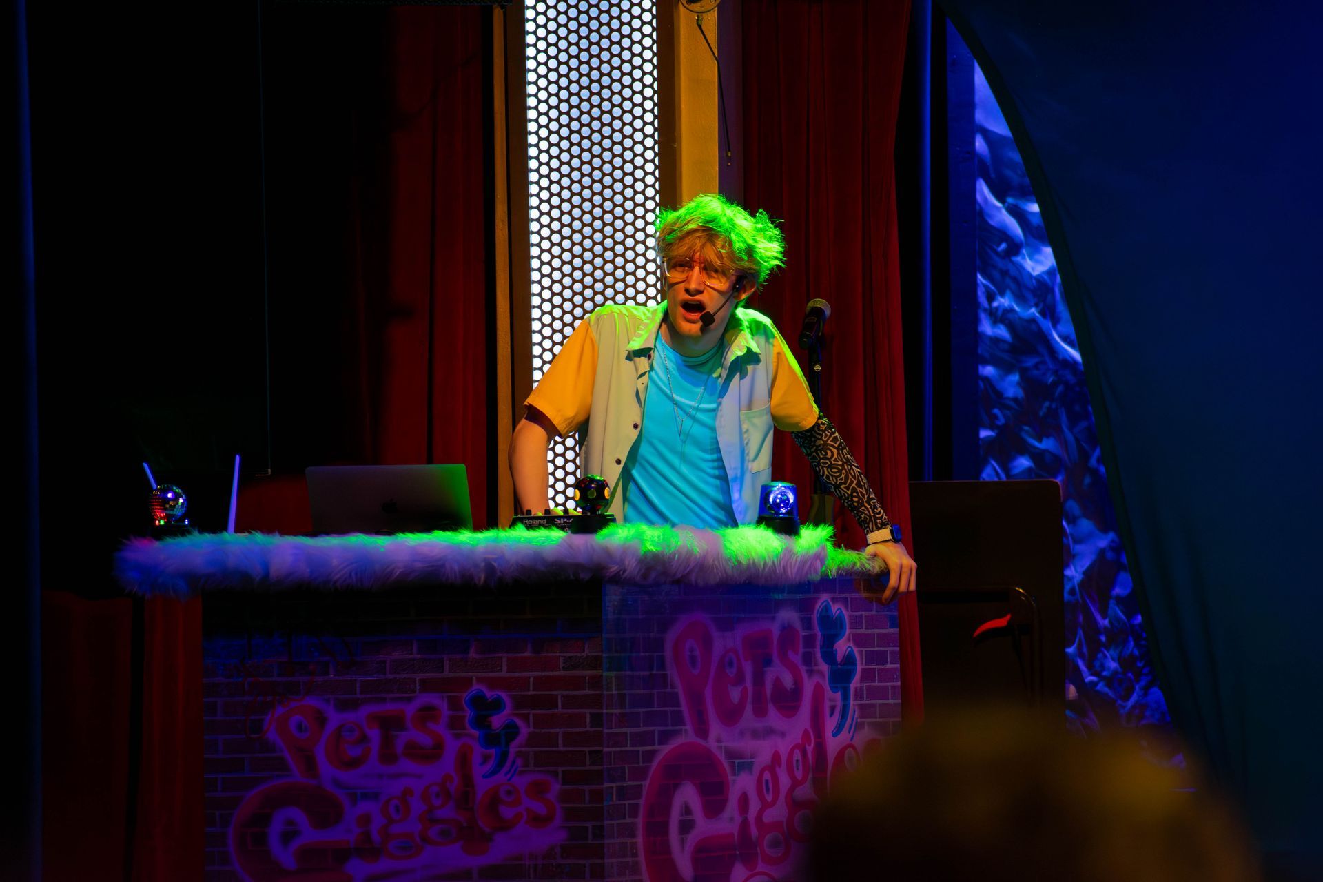 A man in a green wig is standing behind a desk in a dark room.