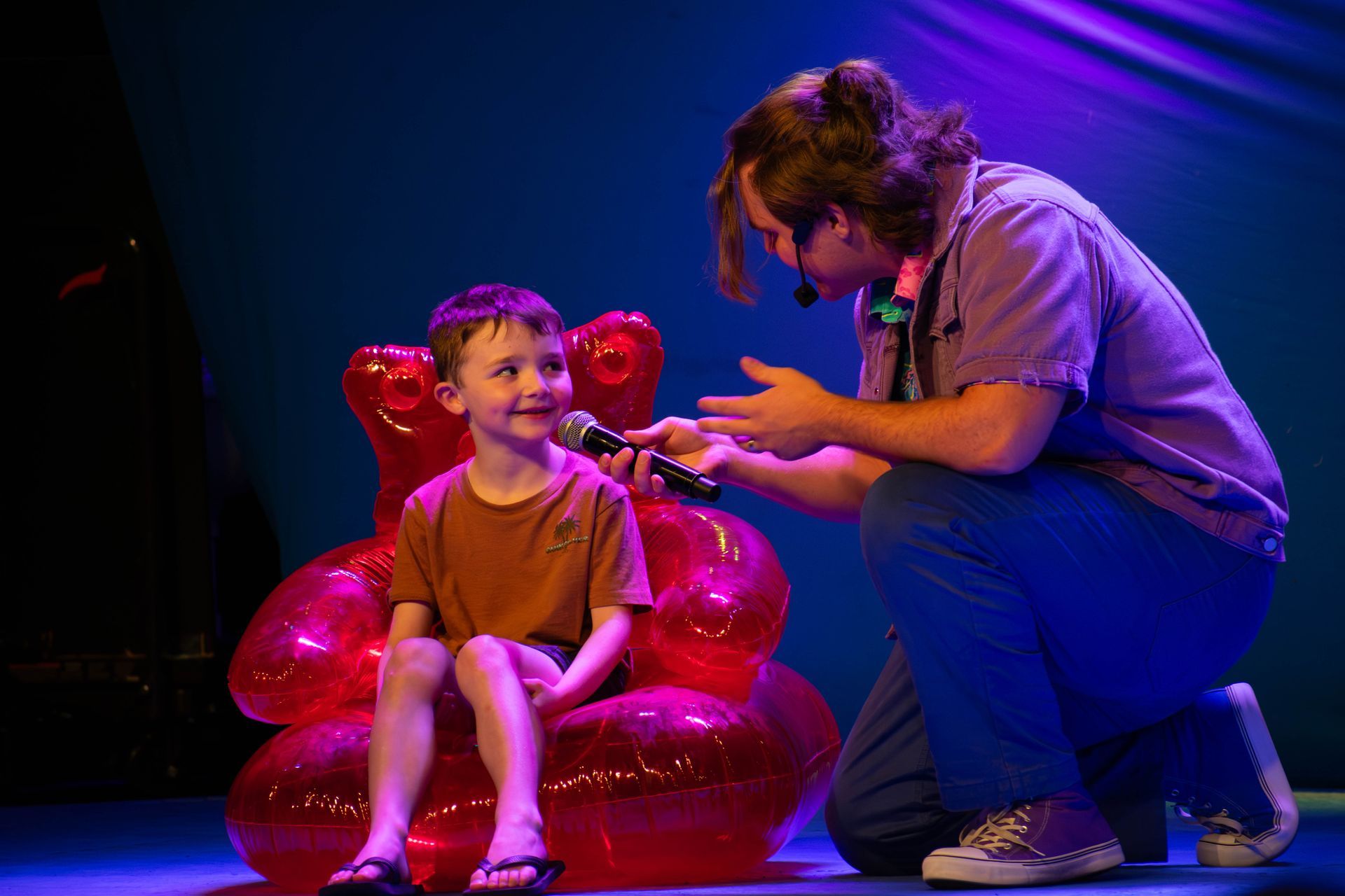 A man is kneeling down next to a young boy sitting in an inflatable chair holding a microphone.
