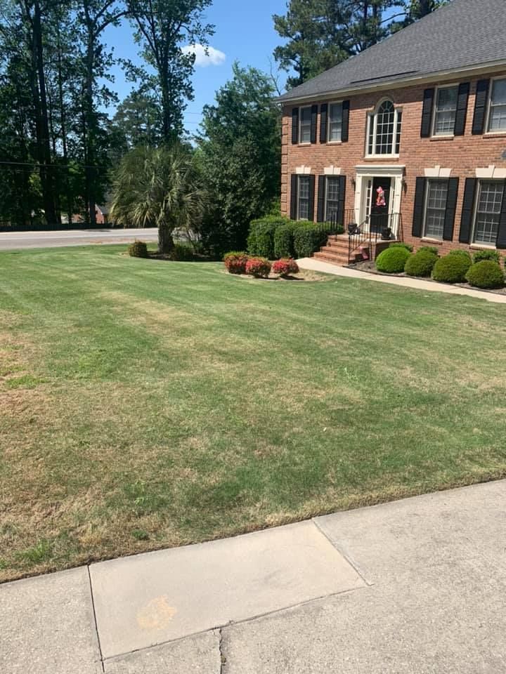 A brick house with a well-manicured lawn, some dry patches, and a sidewalk in front.
