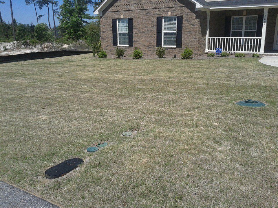 A front yard with brown grass, a brick house, bushes, and utility covers.