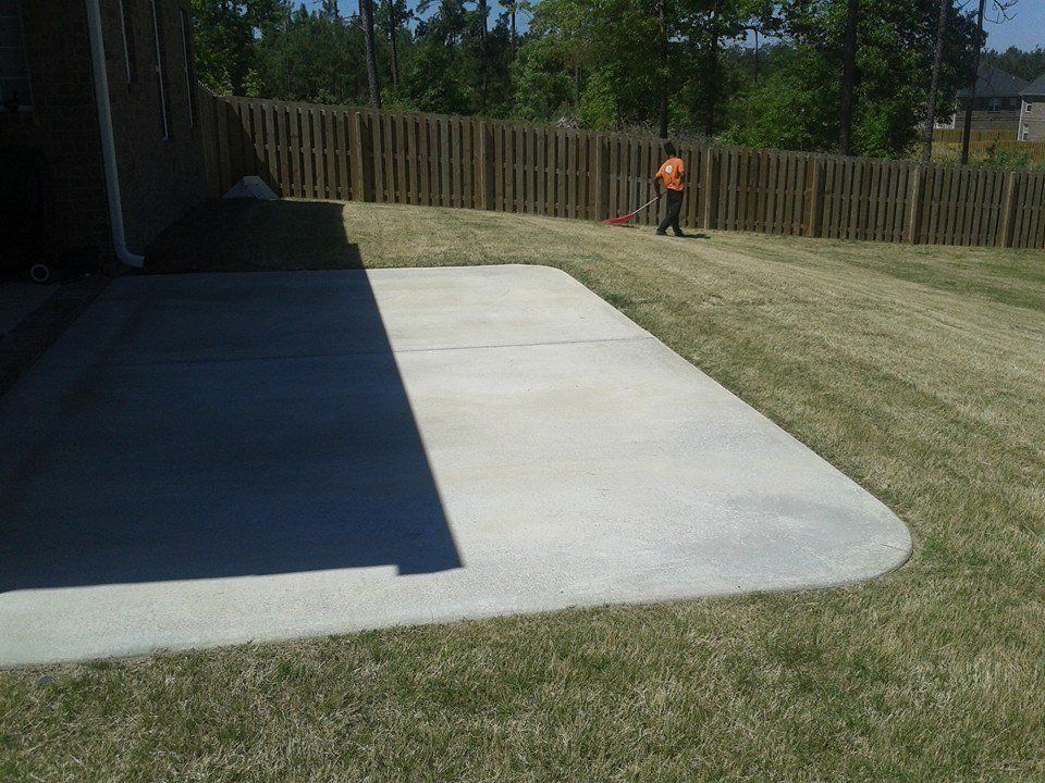 A concrete patio next to a house with a wooden fence, person mowing the grass.