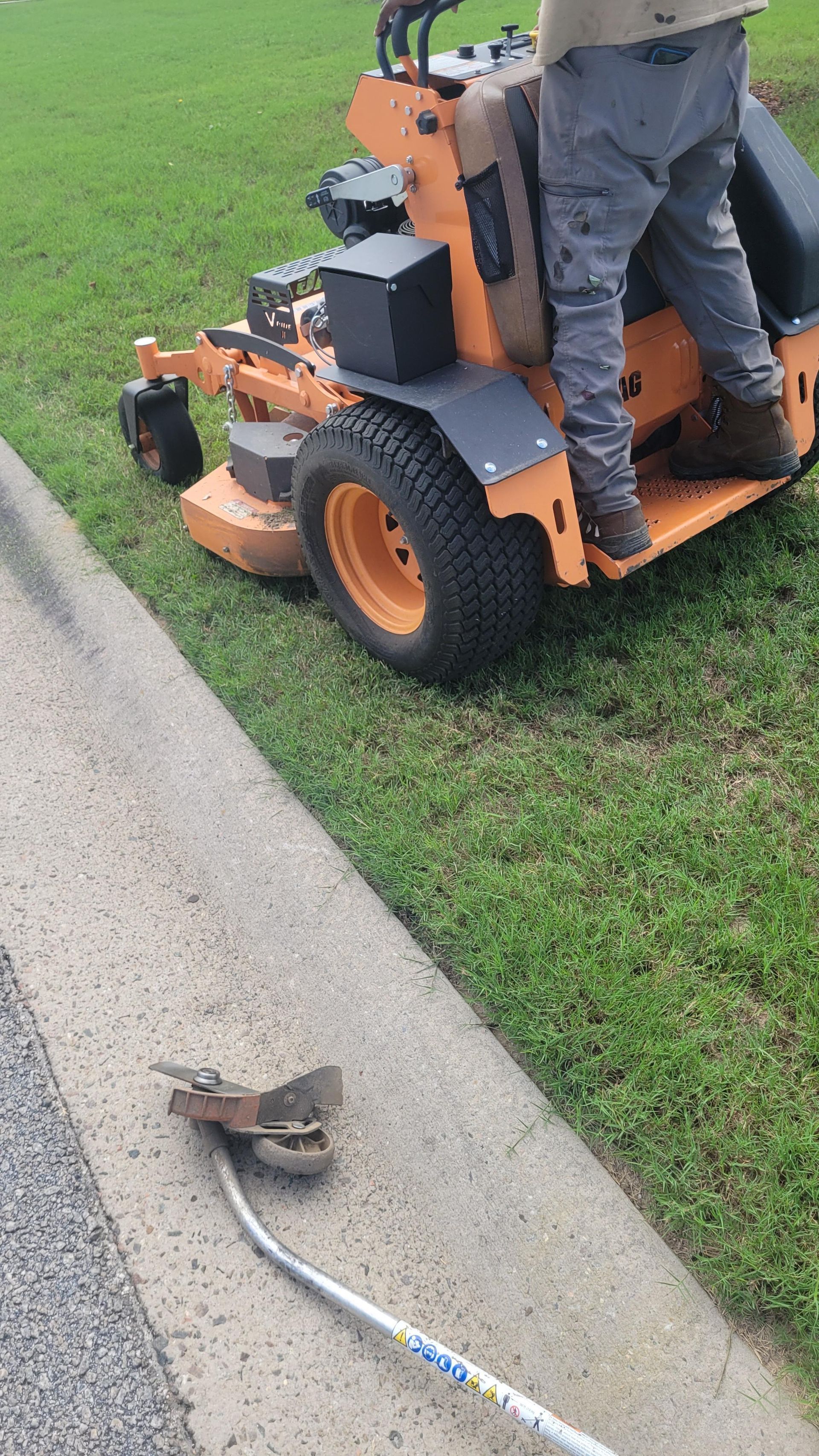 Person operating a riding lawnmower, trimming grass near a curb with a weed wacker on concrete.