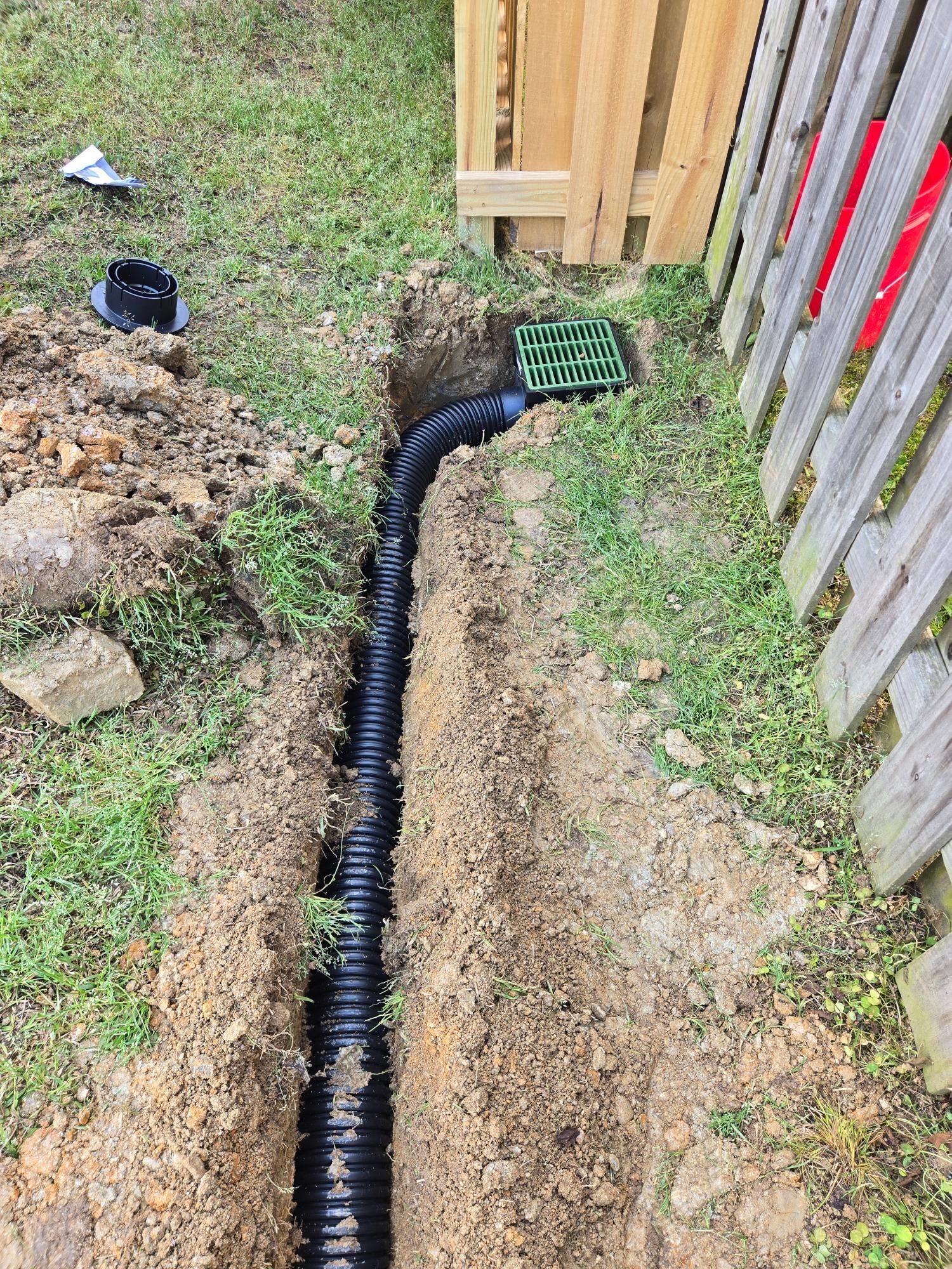 Trench with black drainage pipe running along a fence, green grate installed.