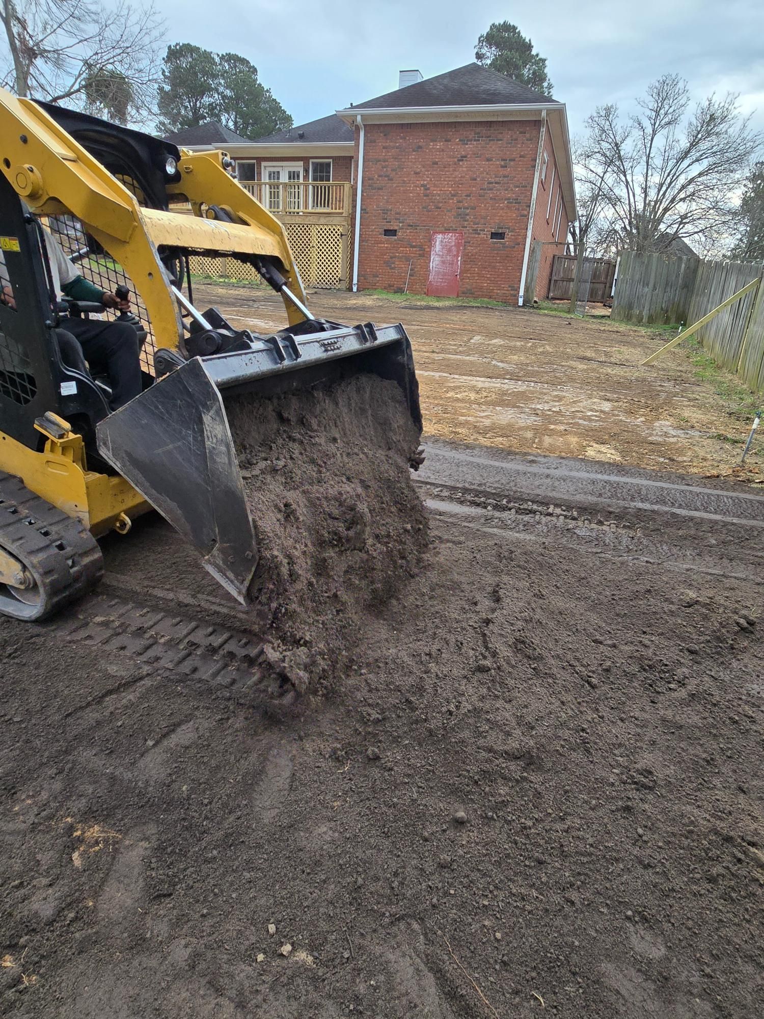 Yellow skid steer dumping soil in a yard. Brown building and cloudy sky in background.