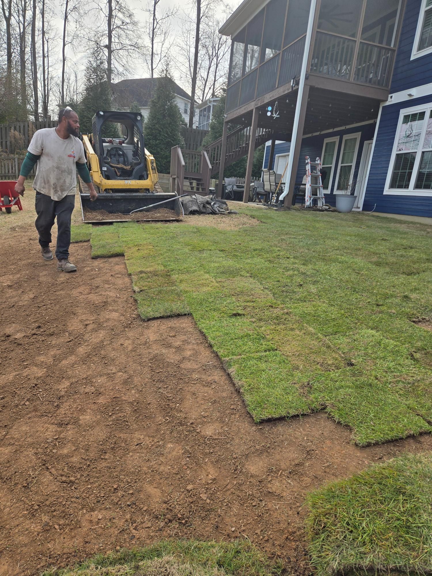 Man laying sod on a residential lawn with a small excavator in the background.