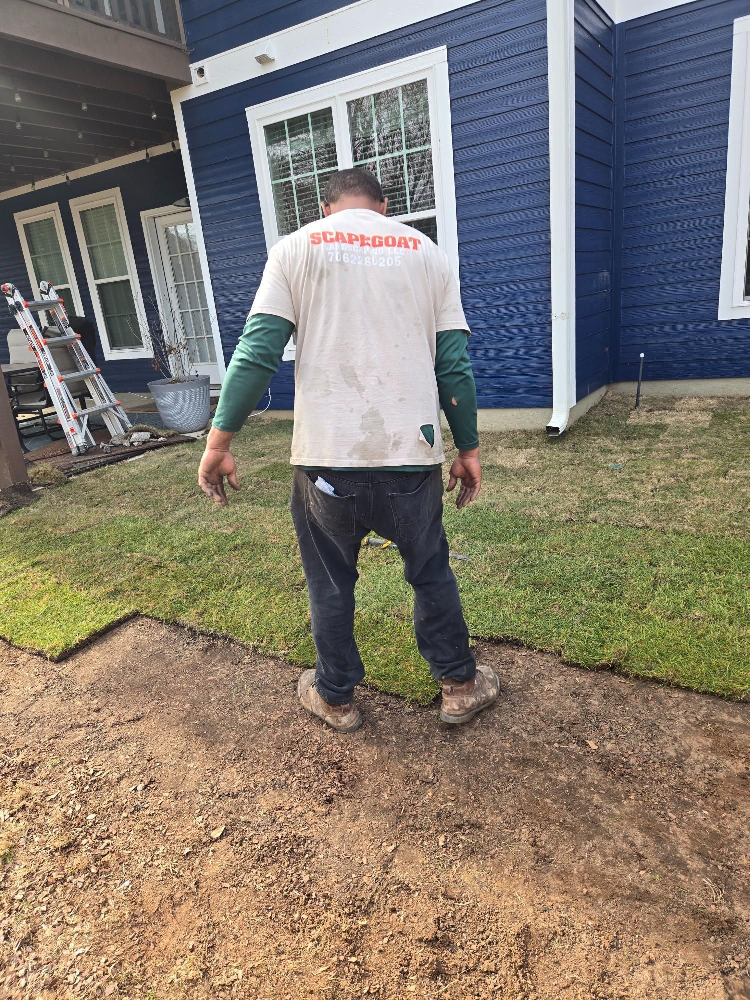 Man in work clothes laying sod near a blue house; outdoors, sunny day.