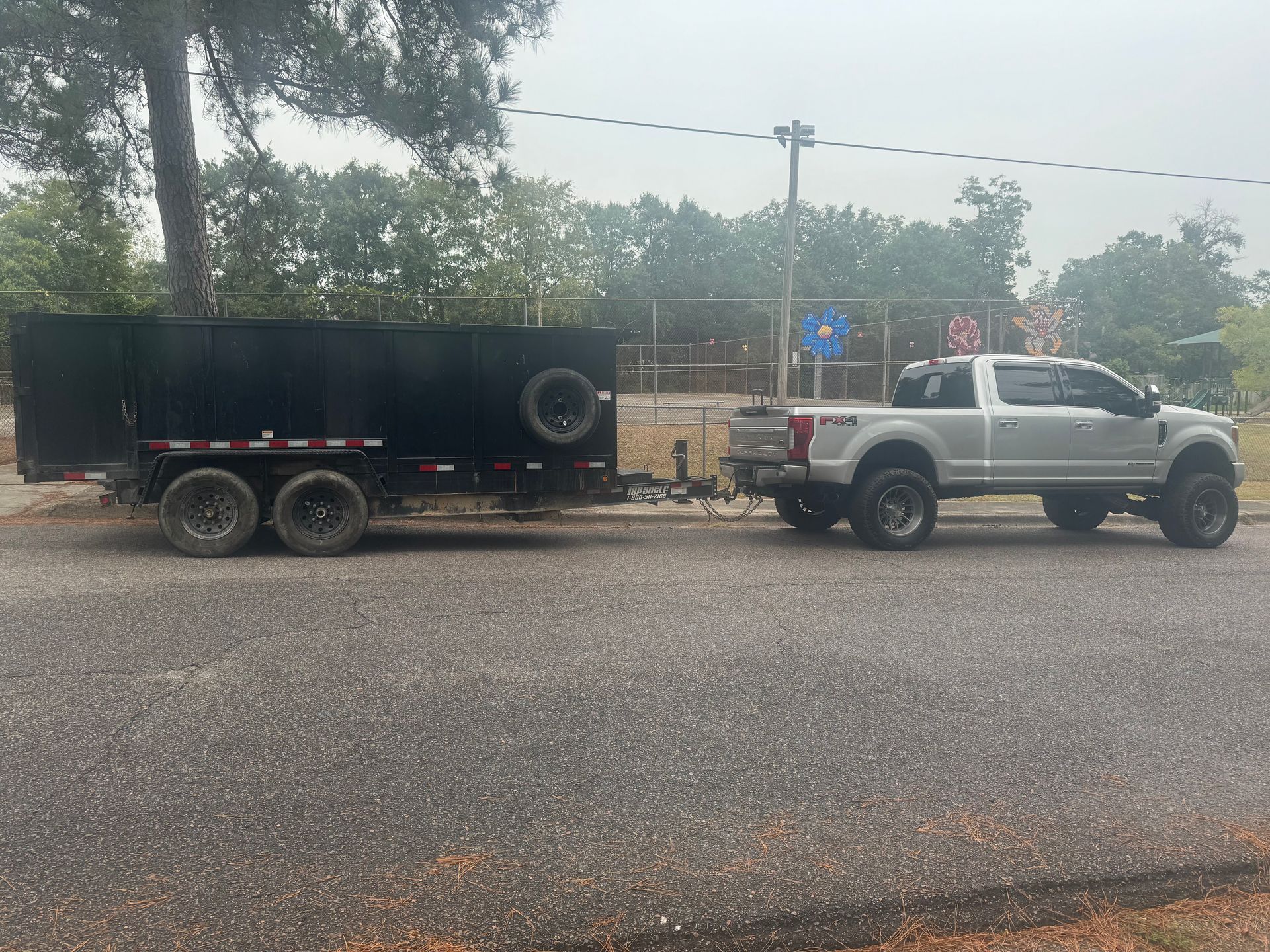 Silver pickup truck towing a black trailer on a road, near a chain-link fence and trees.