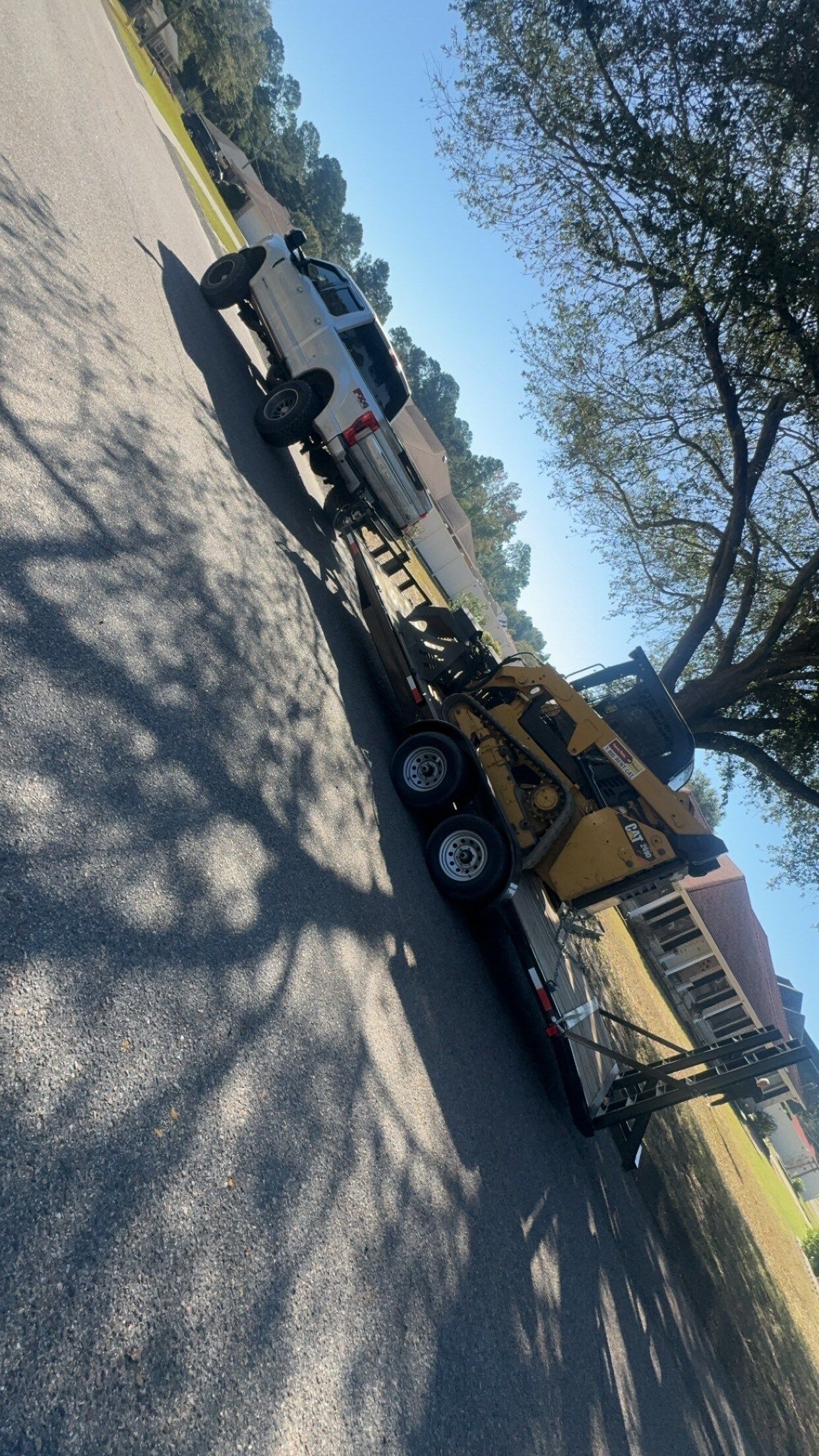 White truck towing a yellow construction machine on a trailer on a paved road under a blue sky.