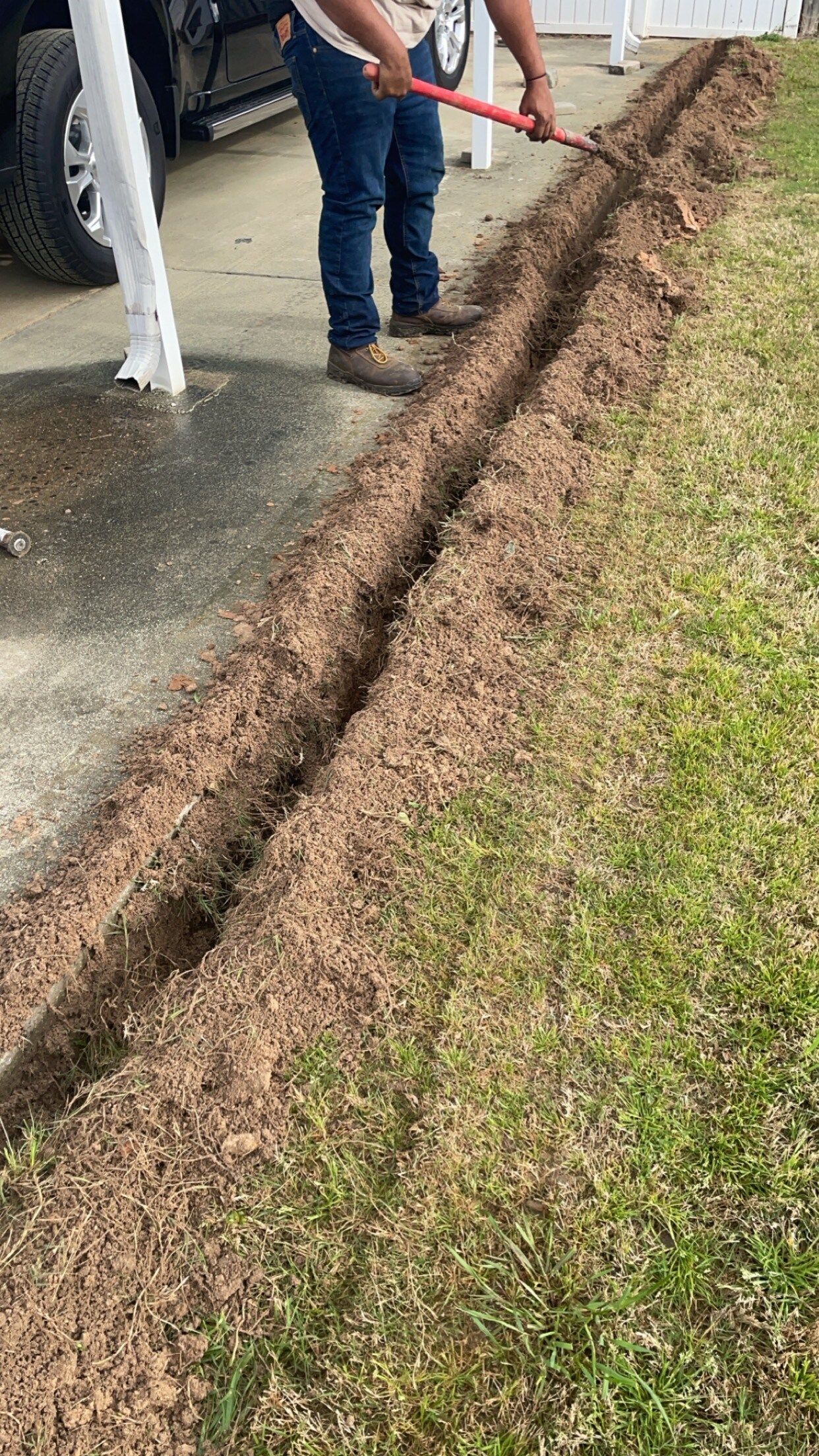 Person digging a narrow trench in grass next to a driveway.