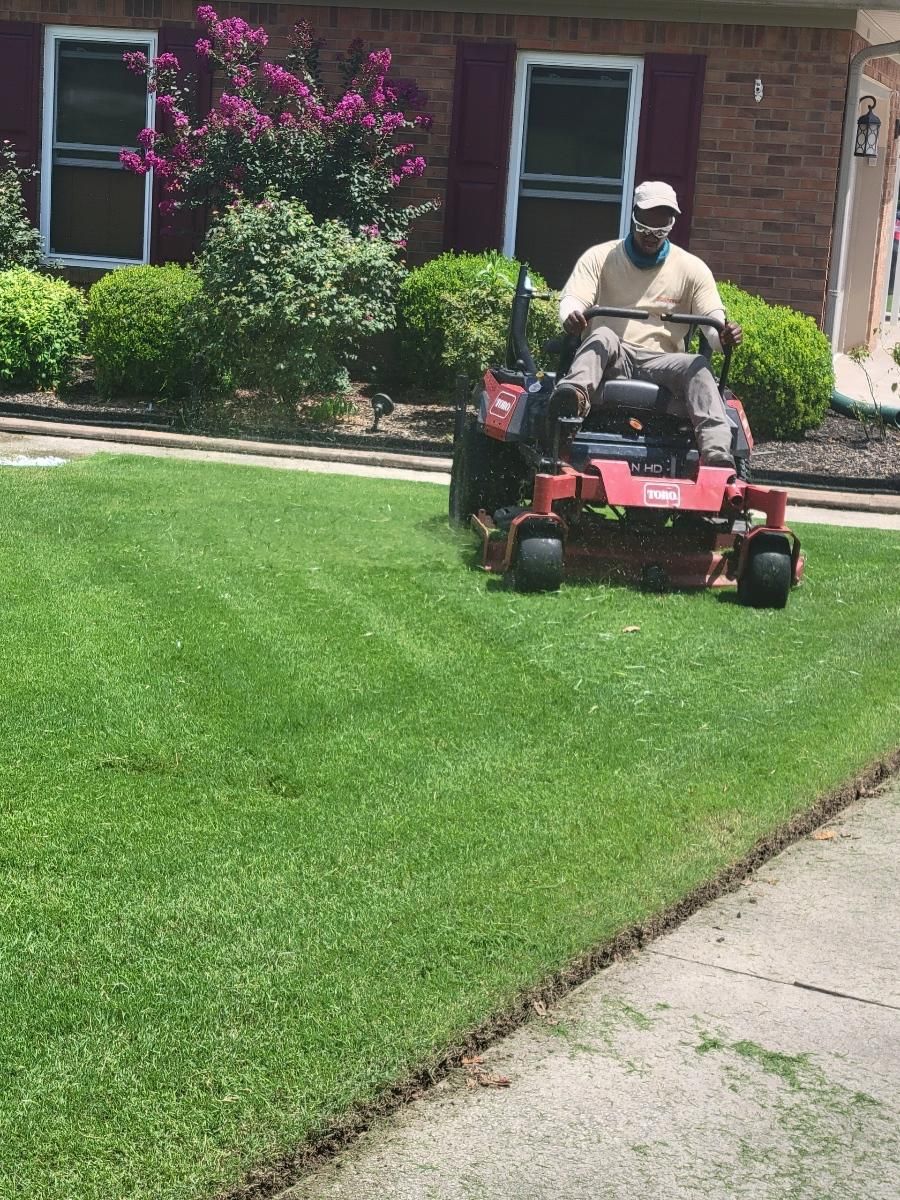 A man mows a green lawn with a red riding lawn mower in front of a house with red shutters and bushes.