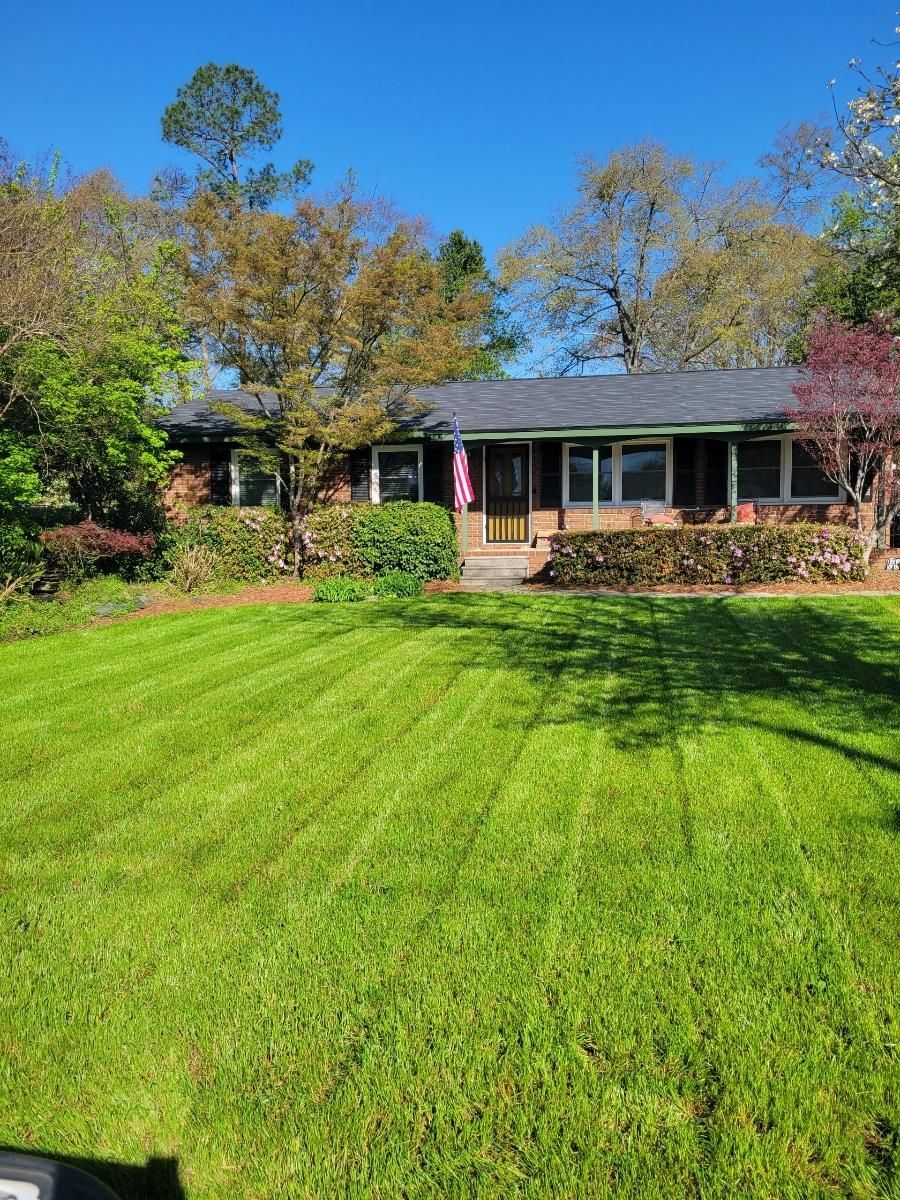 A single-story brick house with a well-manicured lawn, trees, and an American flag on a sunny day.