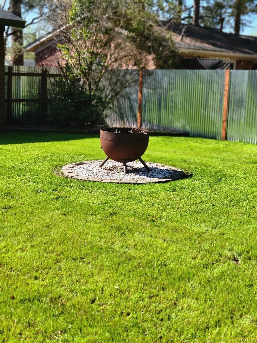 Fire pit on gravel in a grassy backyard with a corrugated metal fence.