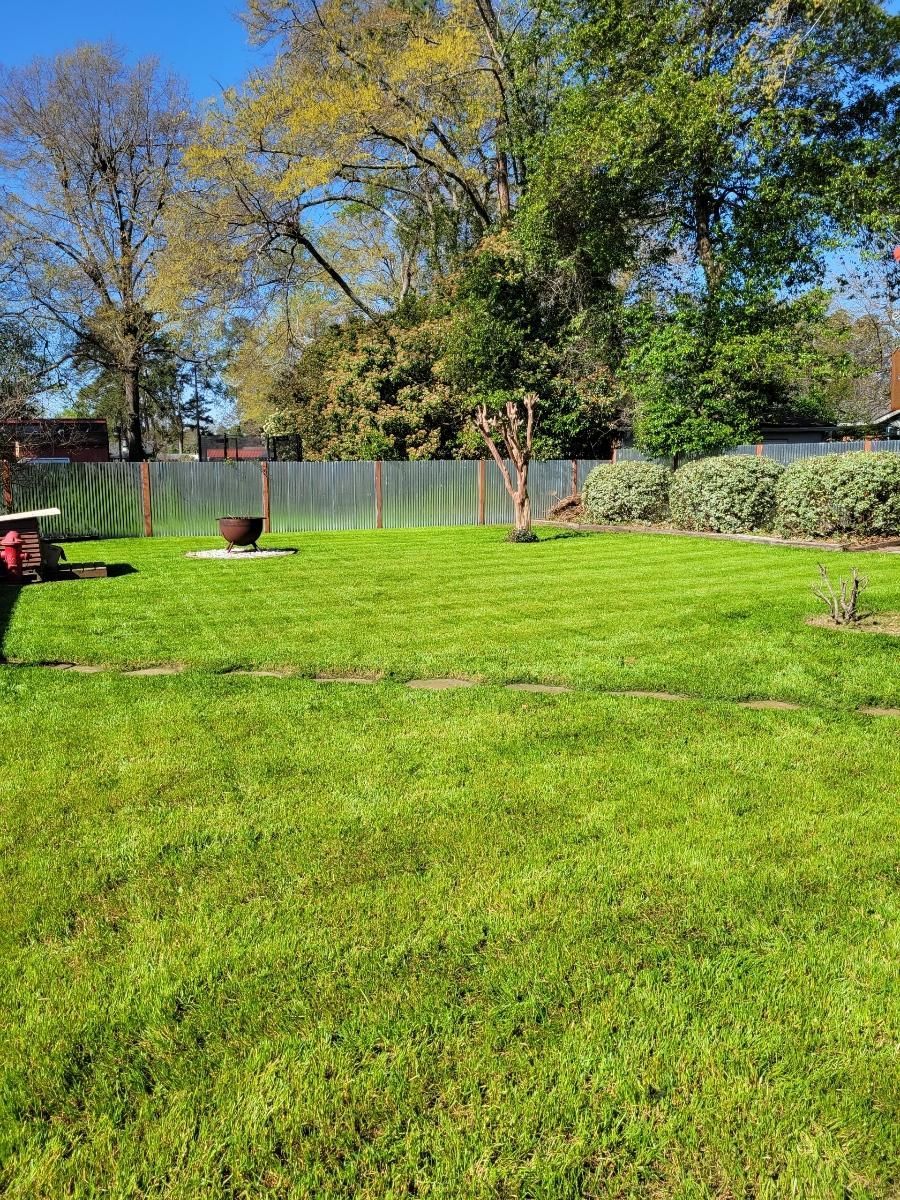 Green lawn in a backyard with a metal fire pit, a fence, trees, and blue sky.