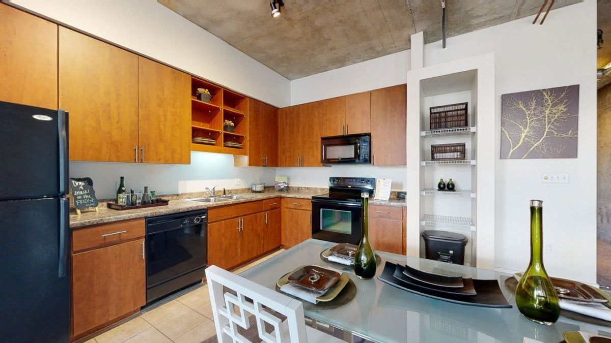 Modern kitchen here at Skyline Lofts Apartments with wood cabinets, granite countertops, and stainless steel appliances. A dining table set with plates and green glass bottles adds elegance.