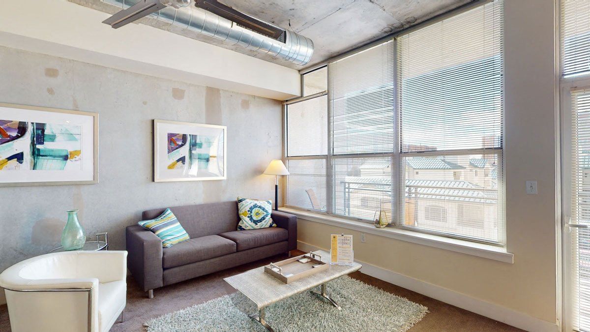Modern living room here at Skyline Lofts Apartments with large windows, gray sofa, colorful cushions, abstract wall art, white armchair, and a glass coffee table on a shag rug.