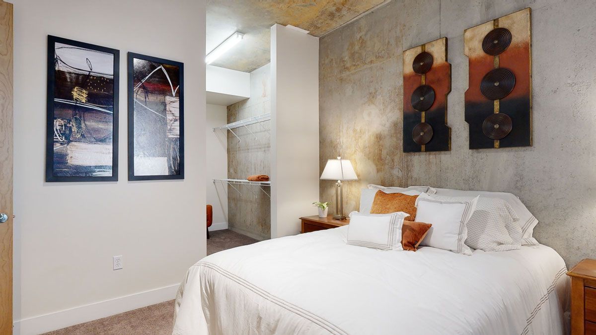 Modern bedroom here at Skyline Lofts Apartments with a cozy white bed, abstract art on concrete walls, a lit lamp on a wooden nightstand, and an open walk-in closet. Calm and inviting.