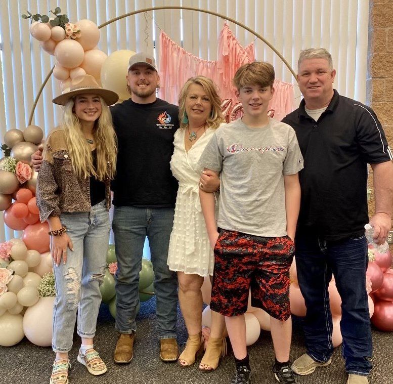 Five people pose in front of a balloon arch. A woman and two others have arms around each other.