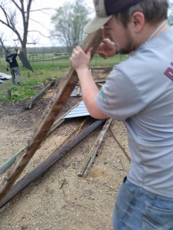 Man in grey shirt, working with wood and metal outside; another person in background.