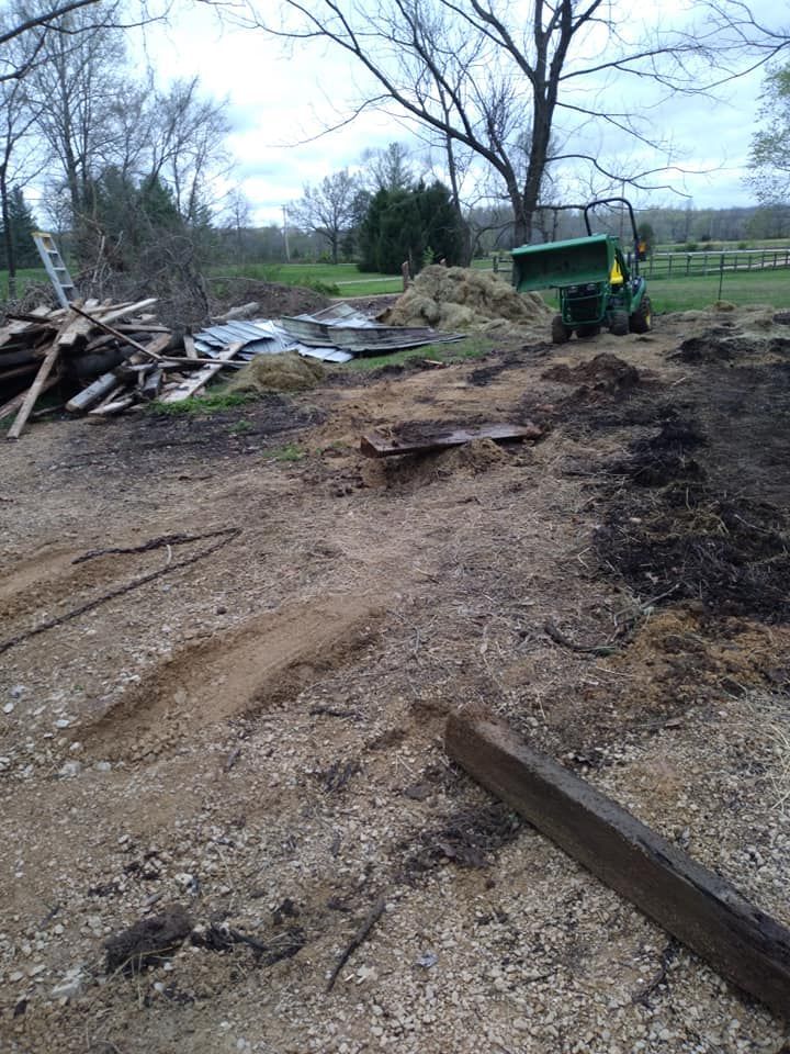 Dirt lot with a green tractor, wood scraps, and a pile of dirt. Trees in the background.